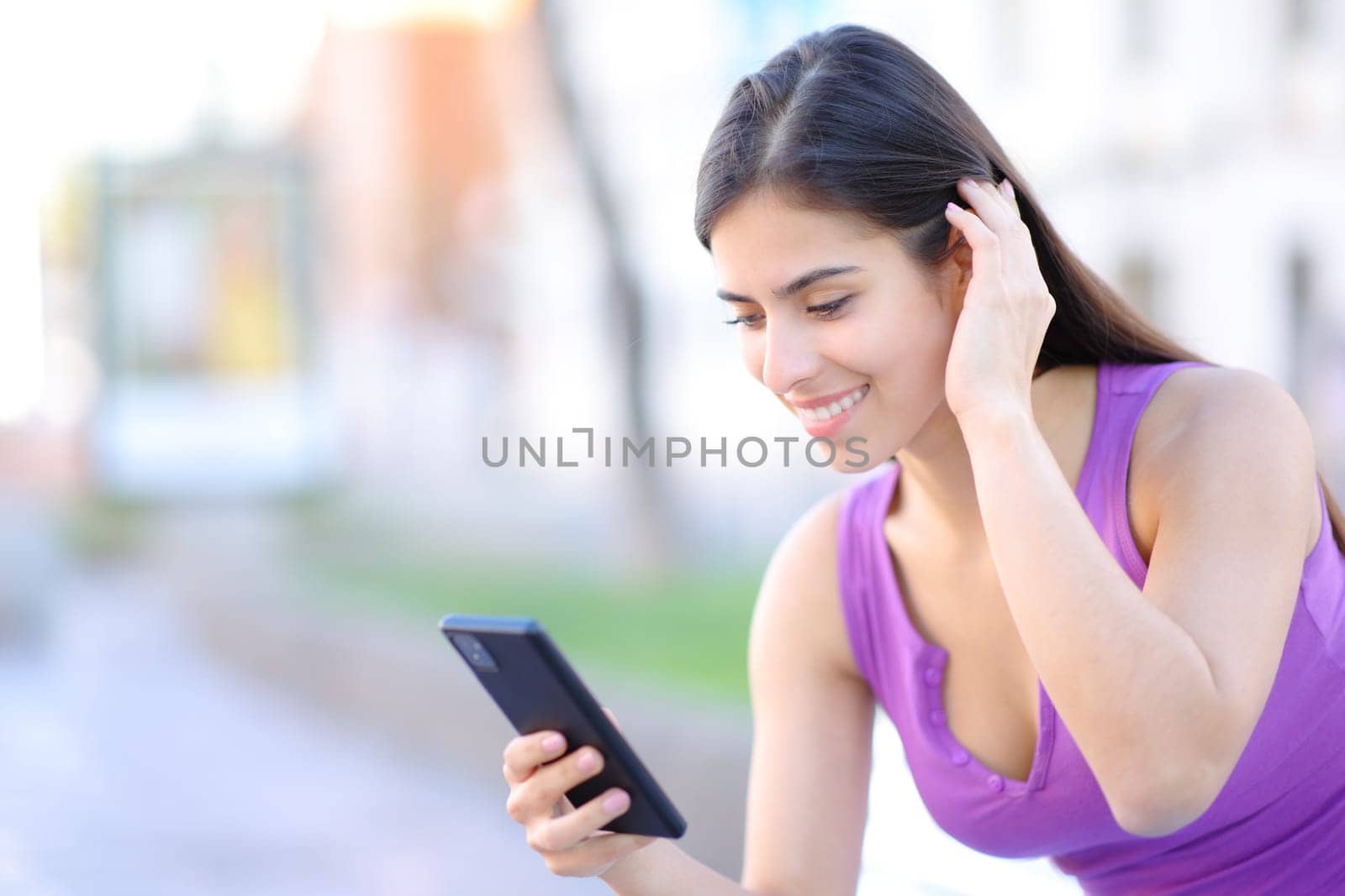 Happy woman using phone touching hair in the street