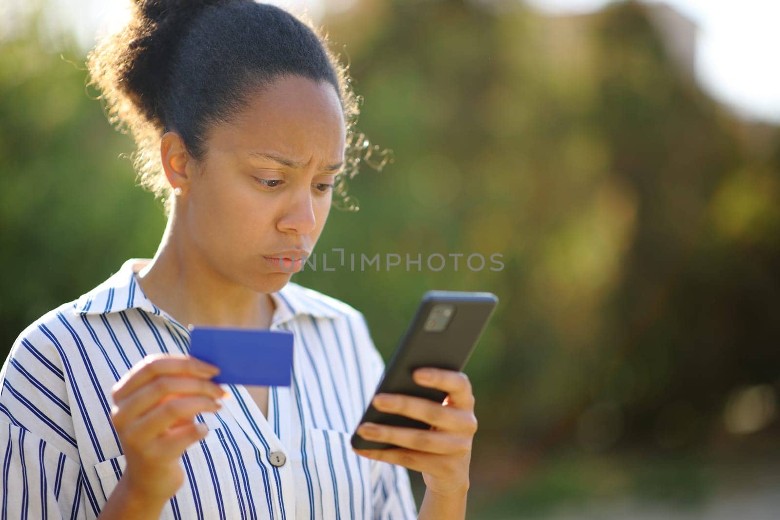 Worried black online buyer holding credit card checking smart phone in a park