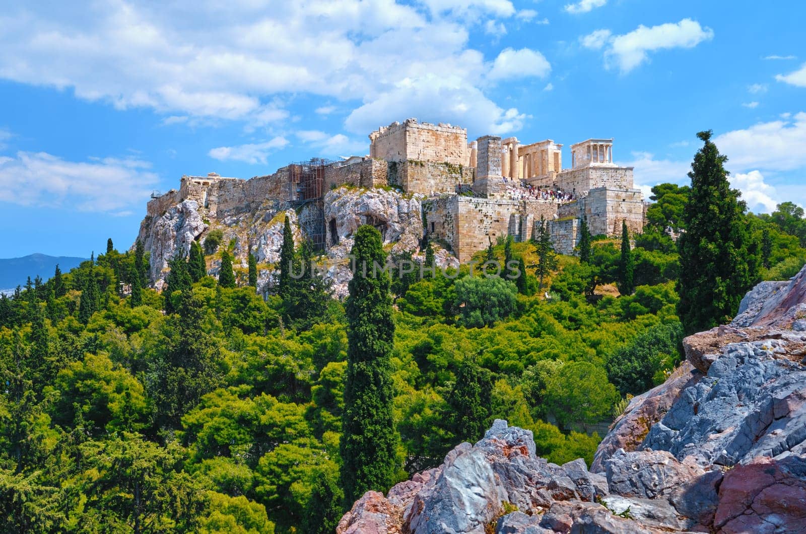 View of Acropolis hill from Areopagus hill in summer daylight with great clouds in blue sky, Athens, Greece. UNESCO world heritage. Propylaea entrance, Agrippa's monument, Parthenon. Fresh green foliage and stones and rocks of hilltop