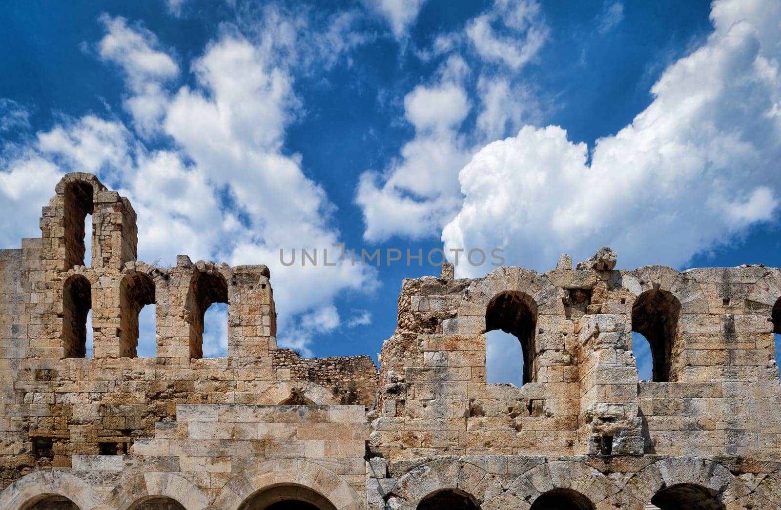 Frontal composition of Odeon of Herodes Atticus theater arches on Acropolis hill, Athens, Greece, against bright blue sky and super clouds. Ancien ruins still used as venue for concerts, performances and festivals. UNESCO world heritage.