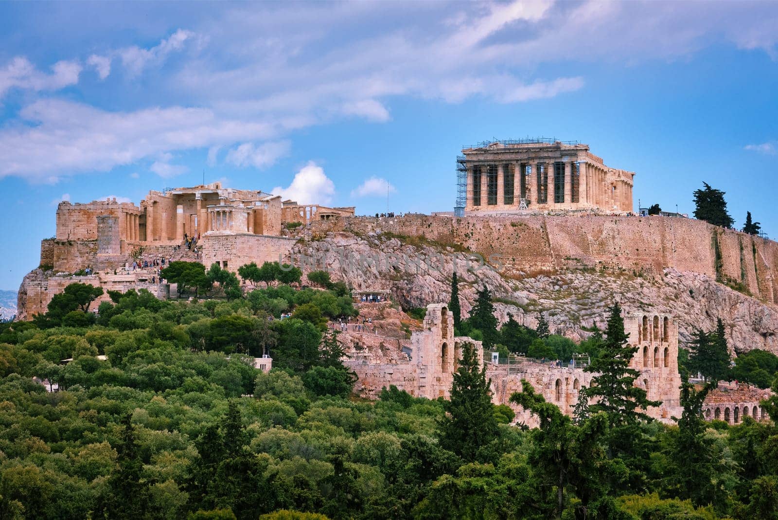 View of Acropolis hill with Propylaea and Parthenon and theater of Odeon below it in Athens, Greece from the hill of Philoppapos or Muses in summer daylight with great clouds in blue sky. Great fresh foliage of olives and cedar trees. Close view