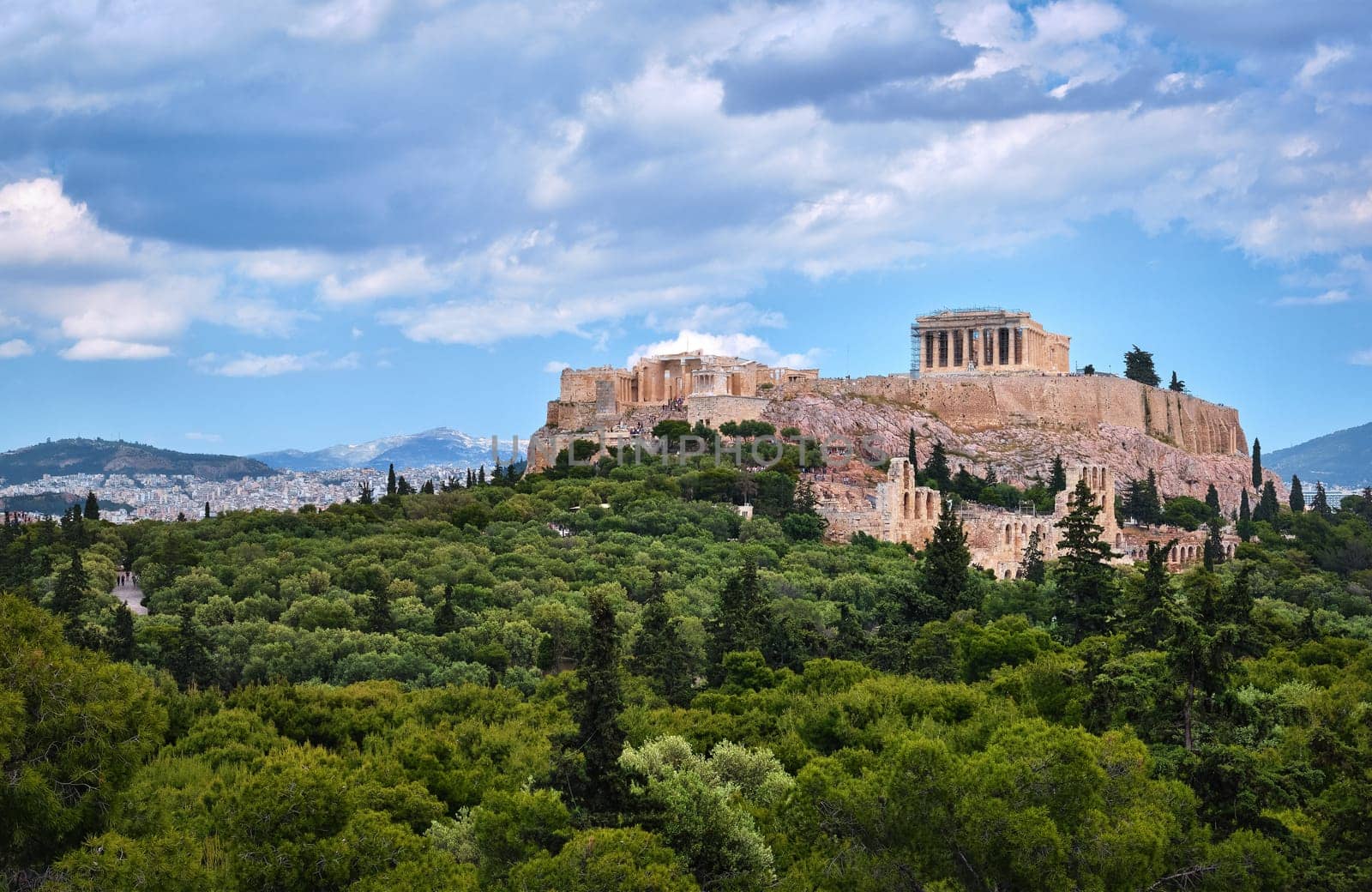 View of Acropolis hill with Propylaea and Parthenon and theater of Odeon below it in Athens, Greece from the hill of Philoppapos or Muses in summer daylight with great clouds in blue sky. Great fresh foliage of olives and cedar trees. Wide shot