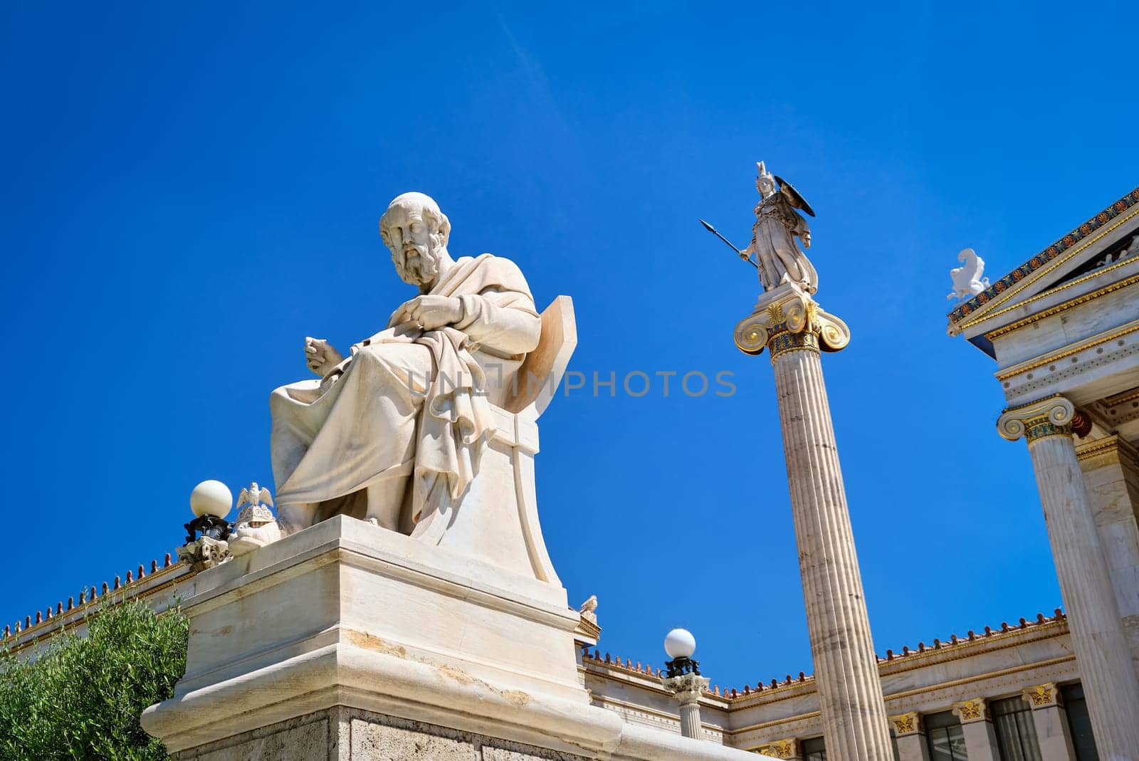 Marble statue of Plato, great ancient Greek philosopher, seated in chair and column or pillar with statue of Athena, ancient Greek goddess, patron of city, in background. Academy of Athens, Athens, Greece. Summer daylight sunshine. Blue sky.