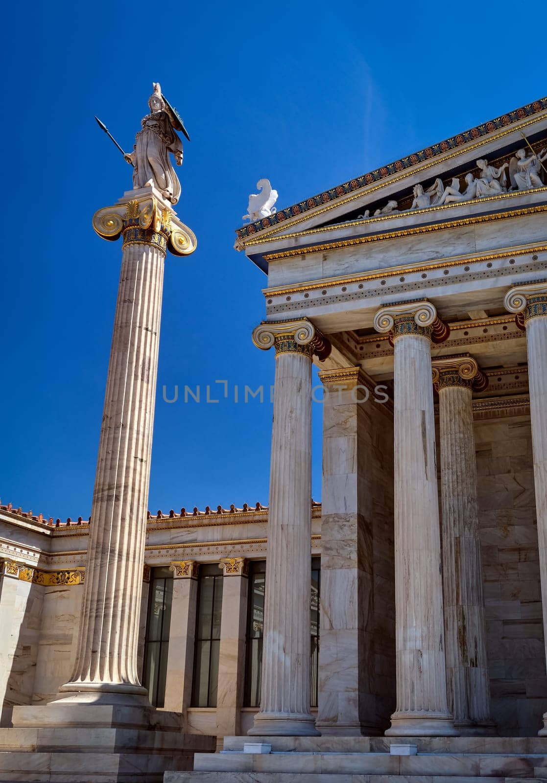 Column or pillar with statue of Athena, ancient Greek goddess, patron of city, by main entrance to Academy of Athens. Athens, Greece, in bright summer day sunshine against blue sky. Vertical shot. Neoclassical architecture.