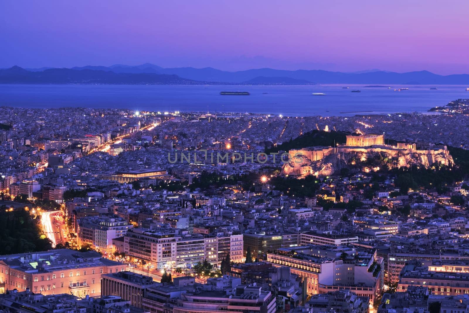 Panoramic wide night view of Athens and Acropolis shot from Lycabettus hill at dusk. Parthenon and Erechtheion lit up by superb night lights. Saronic Gulf and Hellenic Parliament. Famous iconic view of UNESCO world heritage site. After sunset