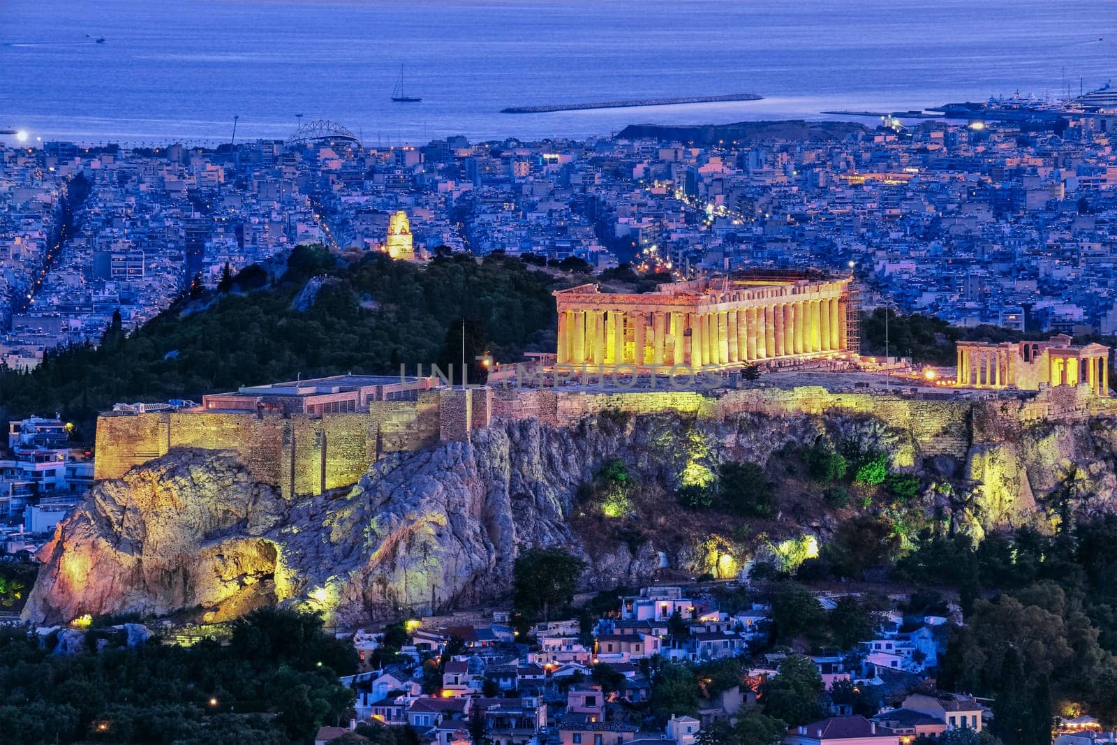 Close view of Acropolis hill with Parthenon and Erechtheion lit up by superb night lights and Philoppapos monument in distance. Night lights of Athens and its harbour. Famous iconic view of UNESCO world heritage site.