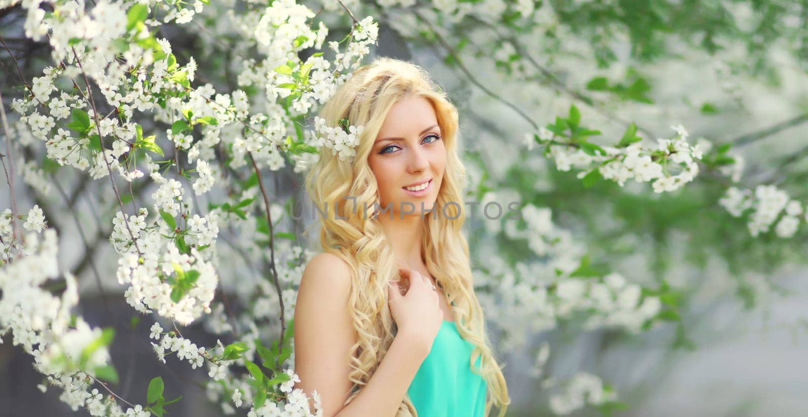 Beautiful young woman with blonde long hair in spring blooming garden on a sunny day against flowers trees background