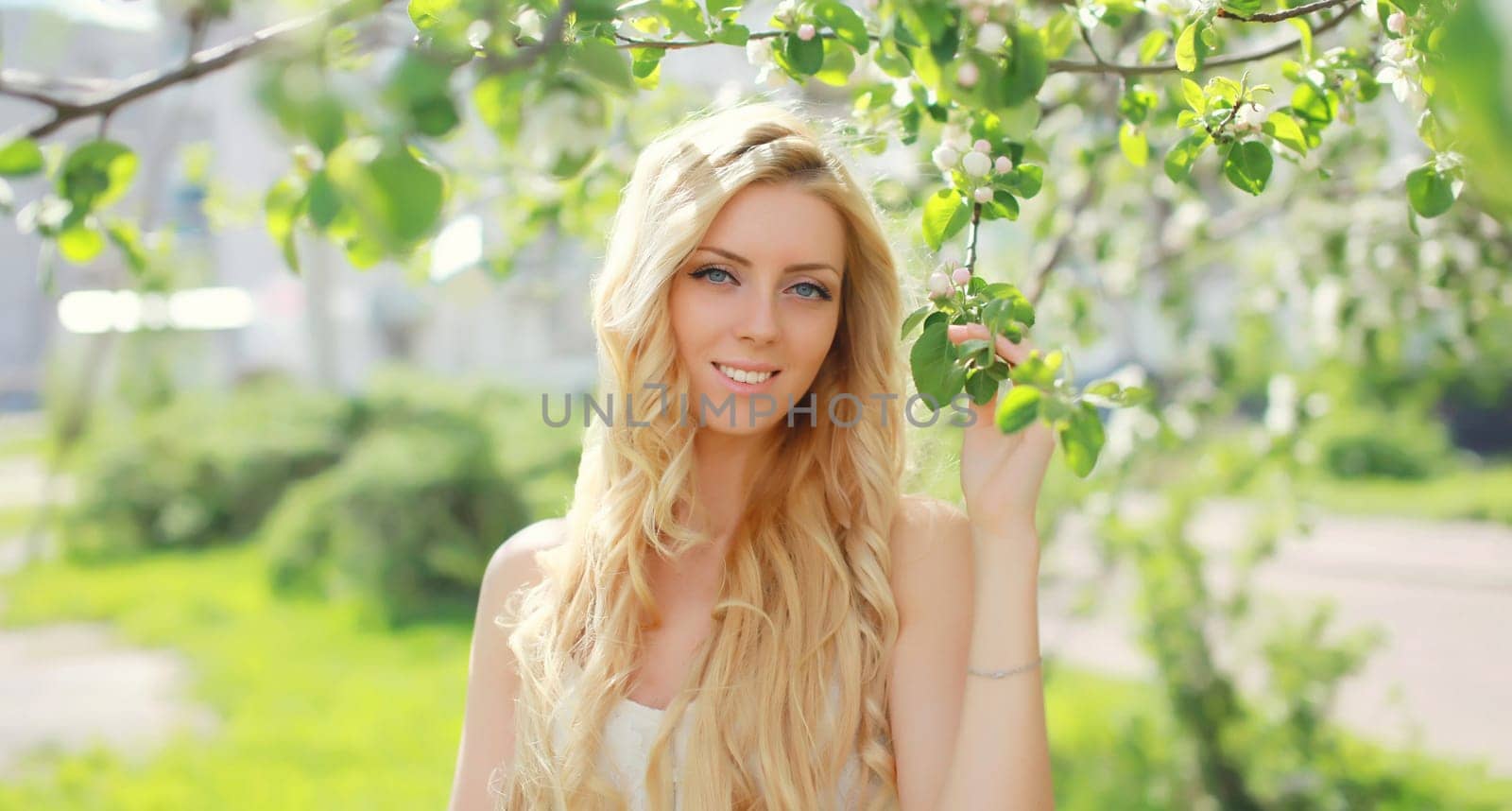 Beautiful young woman with blonde long hair in spring blooming garden on a sunny day against flowers trees background
