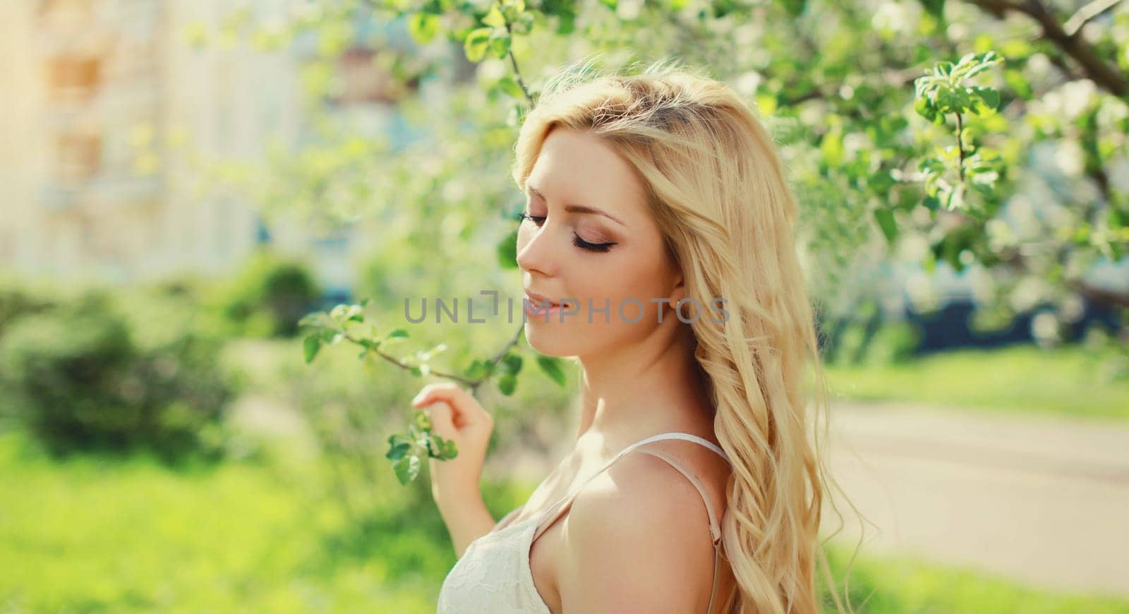 Beautiful young woman with blonde long hair in spring blooming garden on a sunny day against flowers trees background