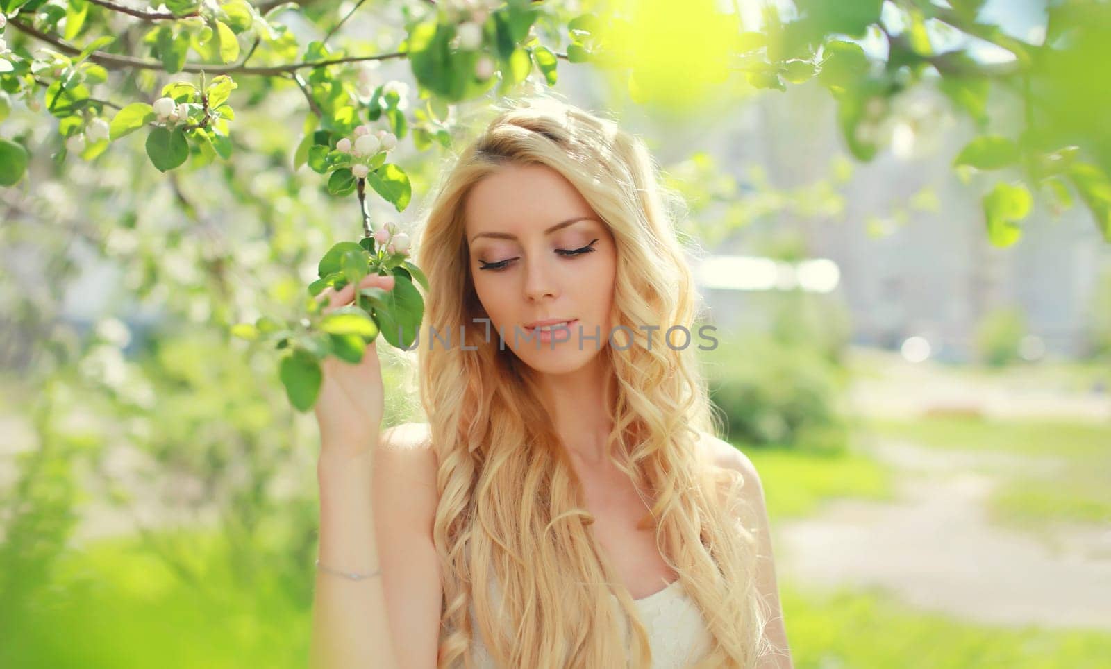 Beautiful young woman with blonde long hair in spring blooming garden on a sunny day against flowers trees background