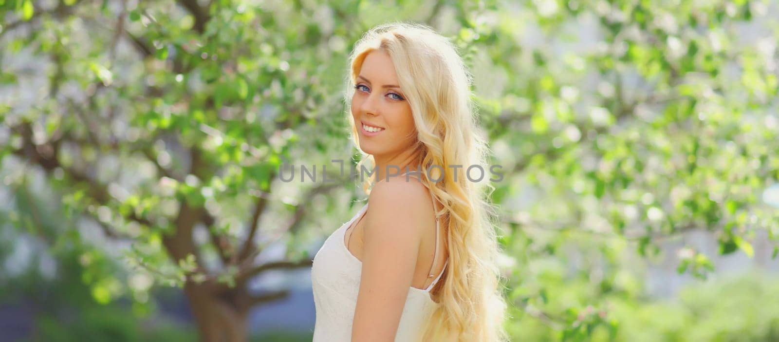 Beautiful young woman with blonde long hair in spring blooming garden on a sunny day against flowers trees background