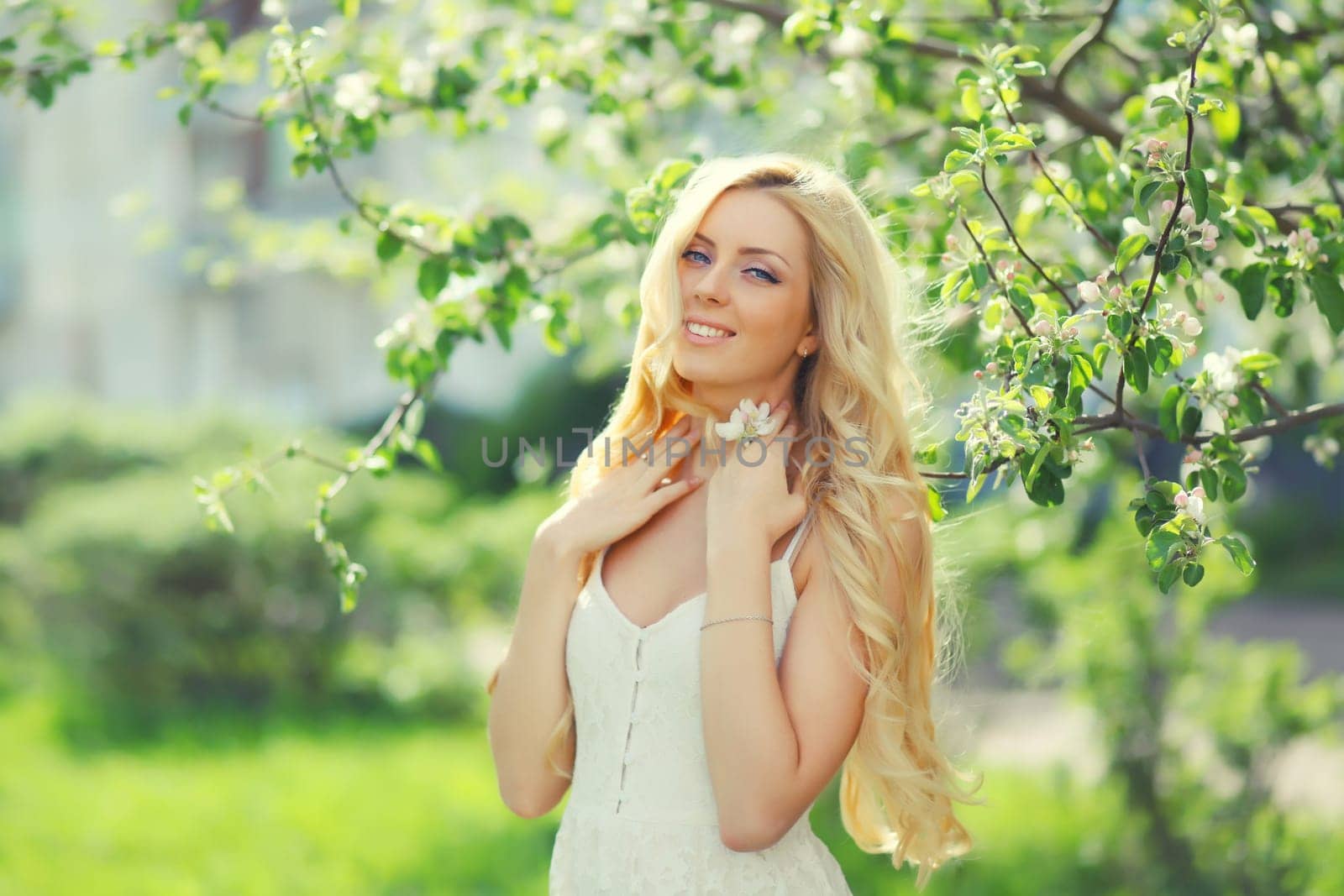 Beautiful young woman with blonde long hair in spring blooming garden on a sunny day against flowers trees background