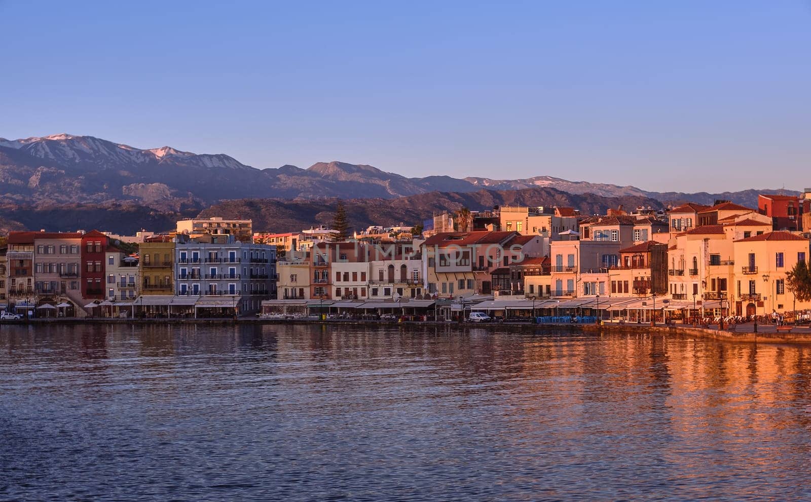 Sunrise view of Old Venetian harbour of Chania, Crete, Greece, shops, hotels, cafes and restaurants on its promenade lit by first sunrays. Cretan hills and mountains in background.