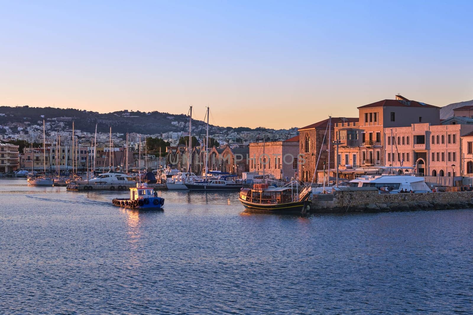 Fishing boat leaving Old Venetian harbour of Chania, Crete, Greece at sunrise. Sailing boats, yachts, tourist cruises anchored by piers. Grand Arsenal and Old Venetian shipyards known as Arsenali Veneziani or Neoria lit by first sunrays.