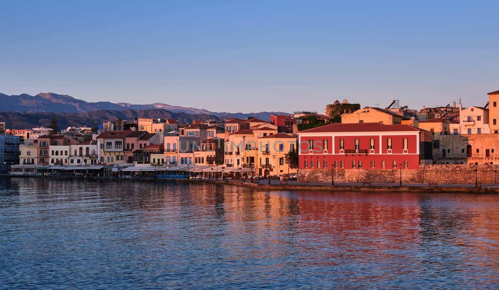 Sunrise view of Old Venetian harbour of Chania, Crete, Greece, shops, hotels, cafes and restaurants on its promenade. Maritime museum of Crete lit by first sunrays. Cretan hills and mountains in background.