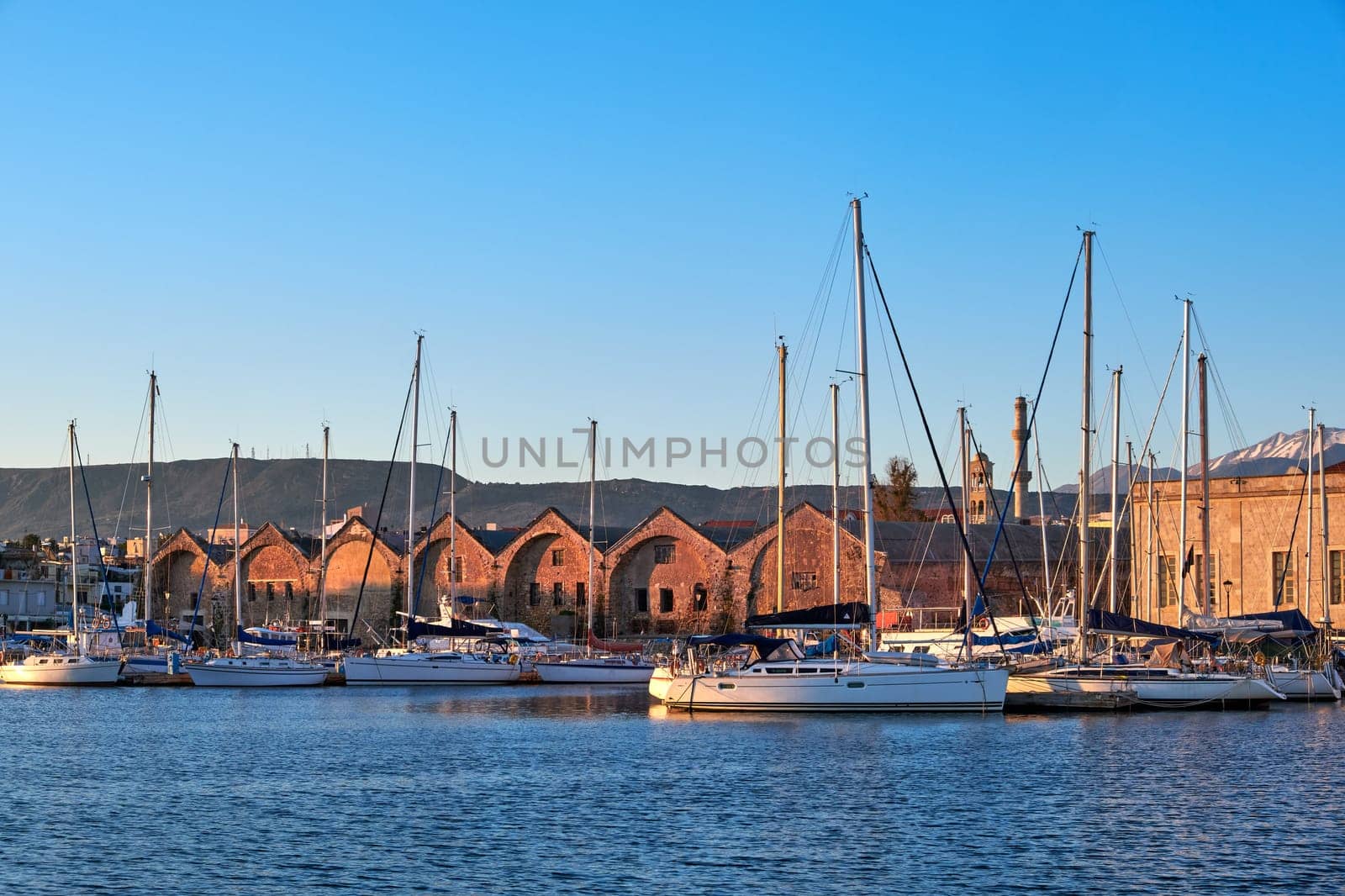 Sunrise over Old Venetian port and harbour of Chania, Crete, Greece. Tourist sailing boats, yachts anchored by pier and Old Venetian shipyards or Arsenali Veneziani or Neoria. Bell tower and minaret of Agios Nikolaos church, distant Cretan mountains