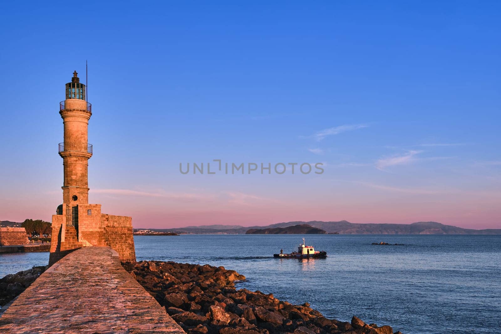 Fishing boat passing by Egyptian Lighthouse lit by first sunrays and leaving Old Venetian harbour of Chania, Crete, Greece at sunrise. Soft early morning colors of sky and Cretan hills and mountains in distance.
