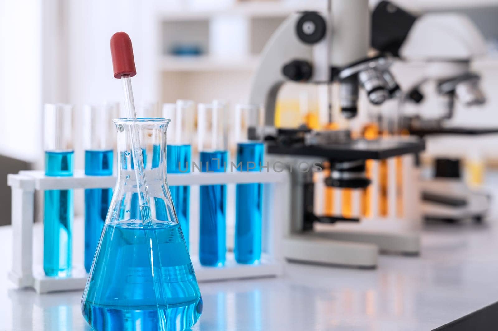 Conical flask glassware with blue solution and dropper, test tubes and microscope in chemical science laboratory in school or university technology background