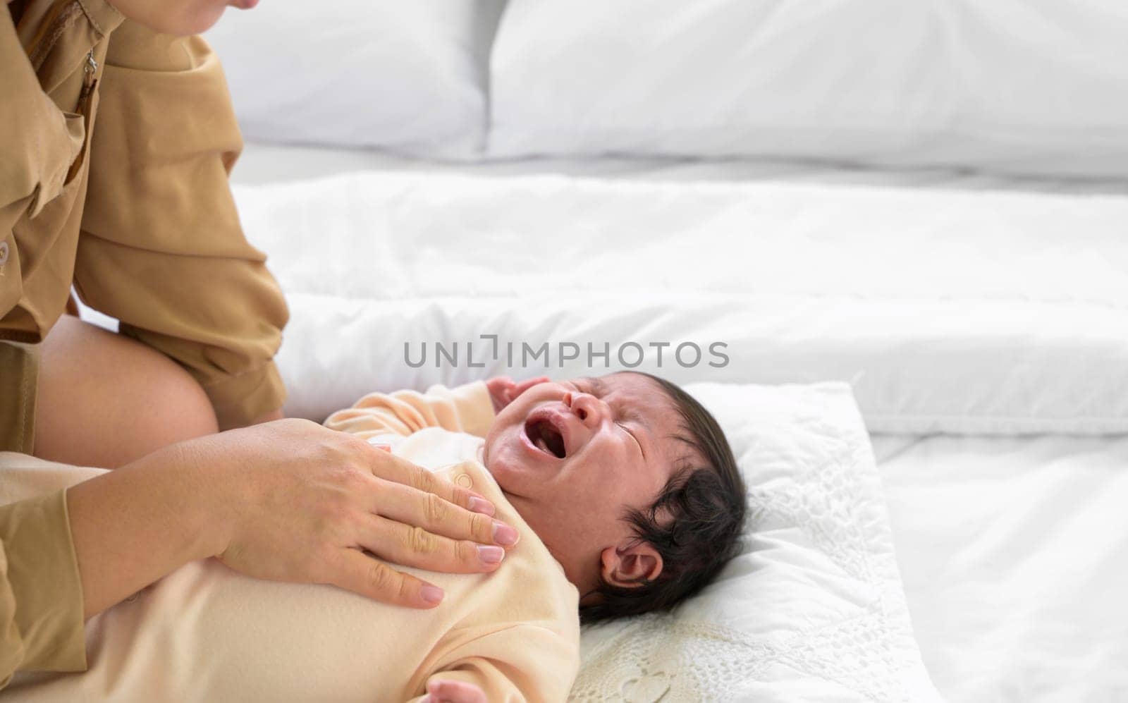 Baby newborn girl crying with mother comforting in bed at home. Mom nursing newborn baby girl and gently touching on chest with love. Healthcare and mother's day