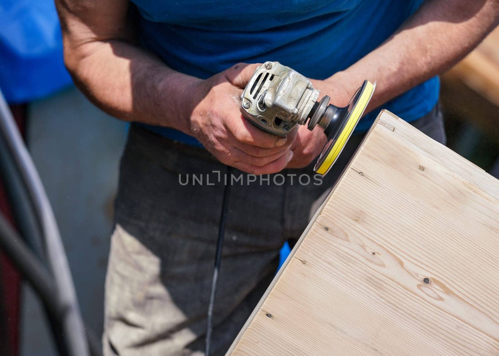 Man polishing wooden chest with old angle grinder during sunny day, closeup detail to hands without gloves.