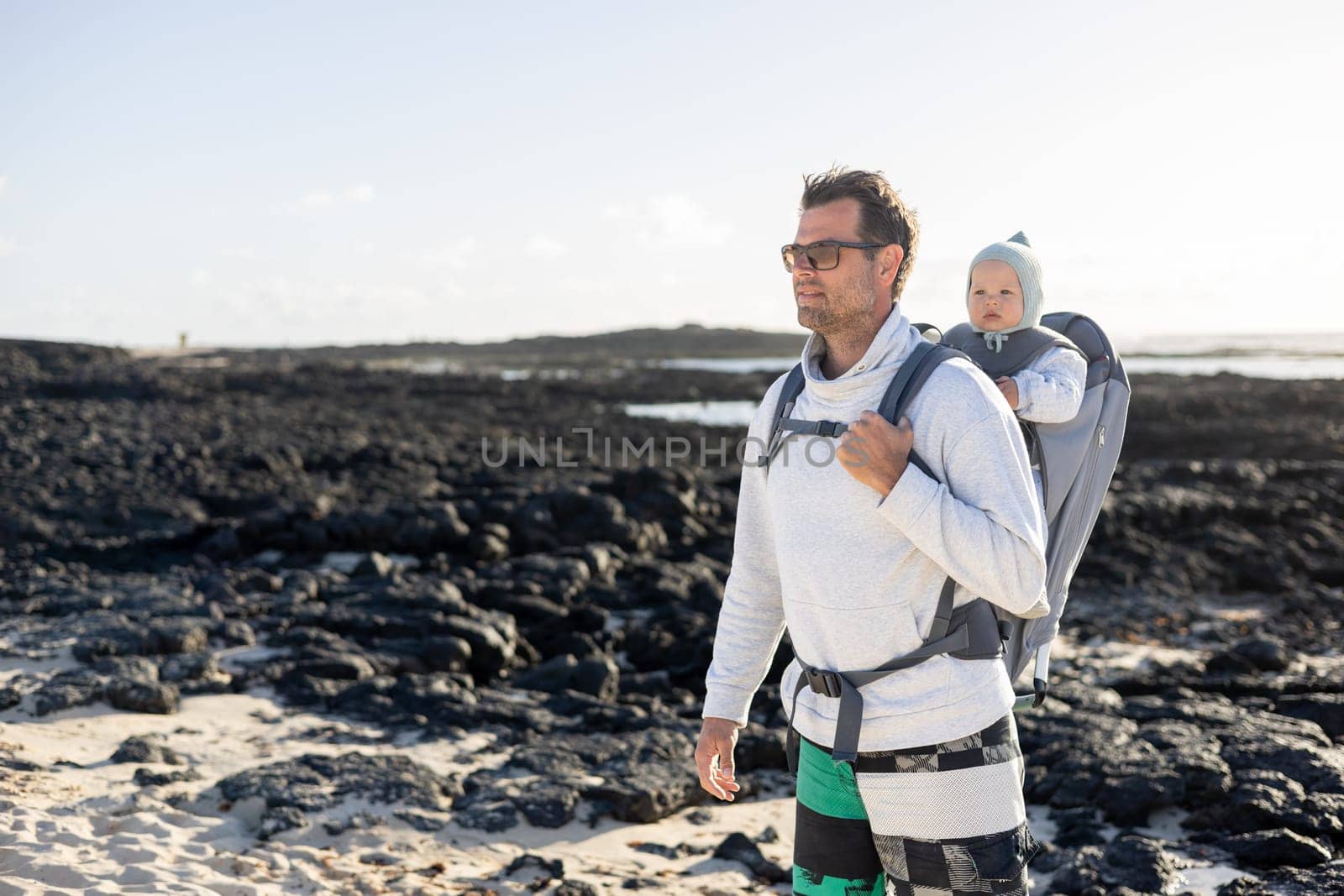 Young father carrying his infant baby boy son in backpack on black rock volcanic beach on Lanzarote island, Spain. Family travel and winter vacation concept