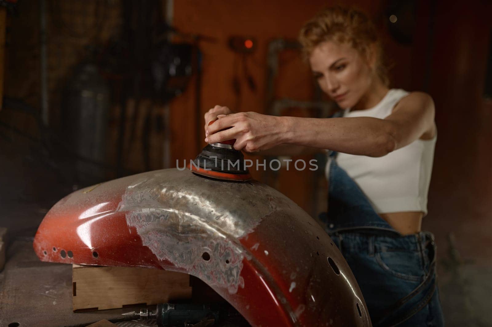 Young woman doing hard job at motorcycle garage using grinder to repair some metal bike part