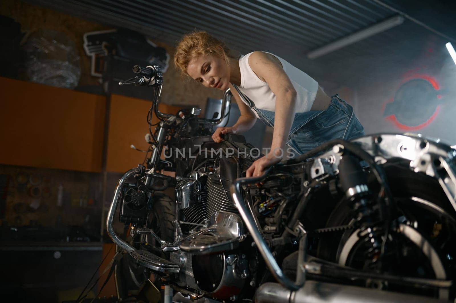 Beautiful woman mechanic posing for camera nearby motorcycle at workshop. Young female technician after hard job in garage service