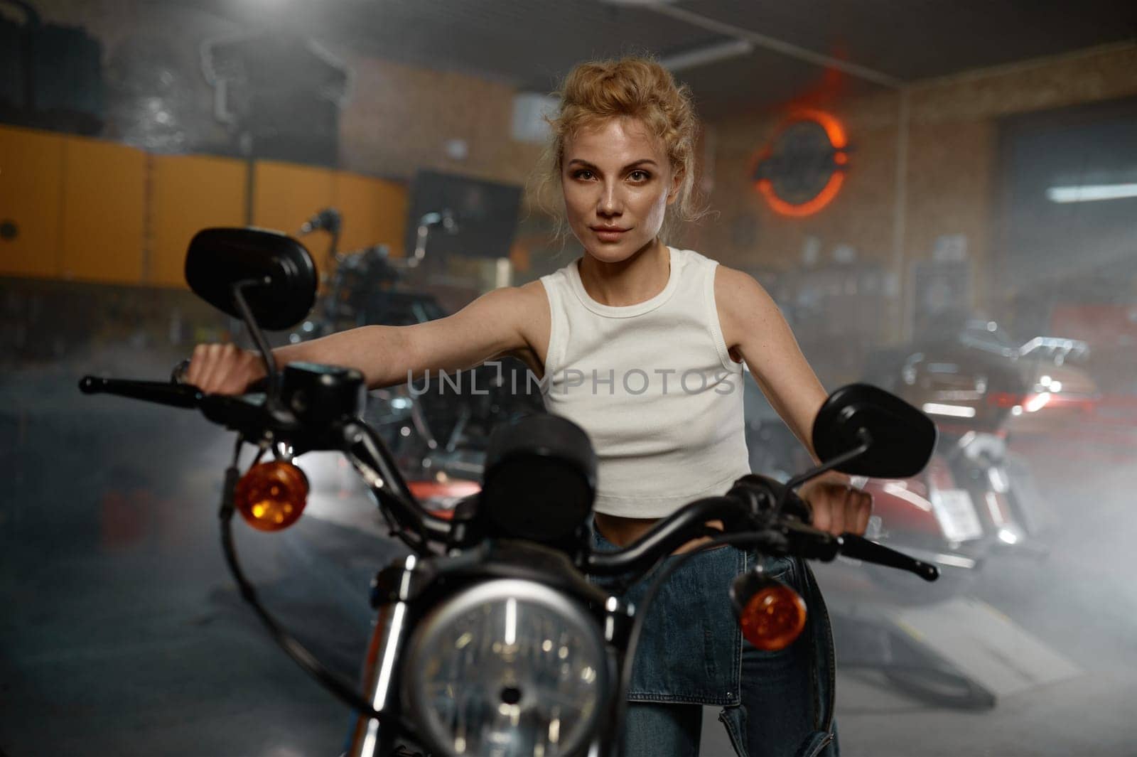 Young woman motorcyclist testing repaired custom sportbike. Indoor portrait headshot of female biker or mechanic over garage interior