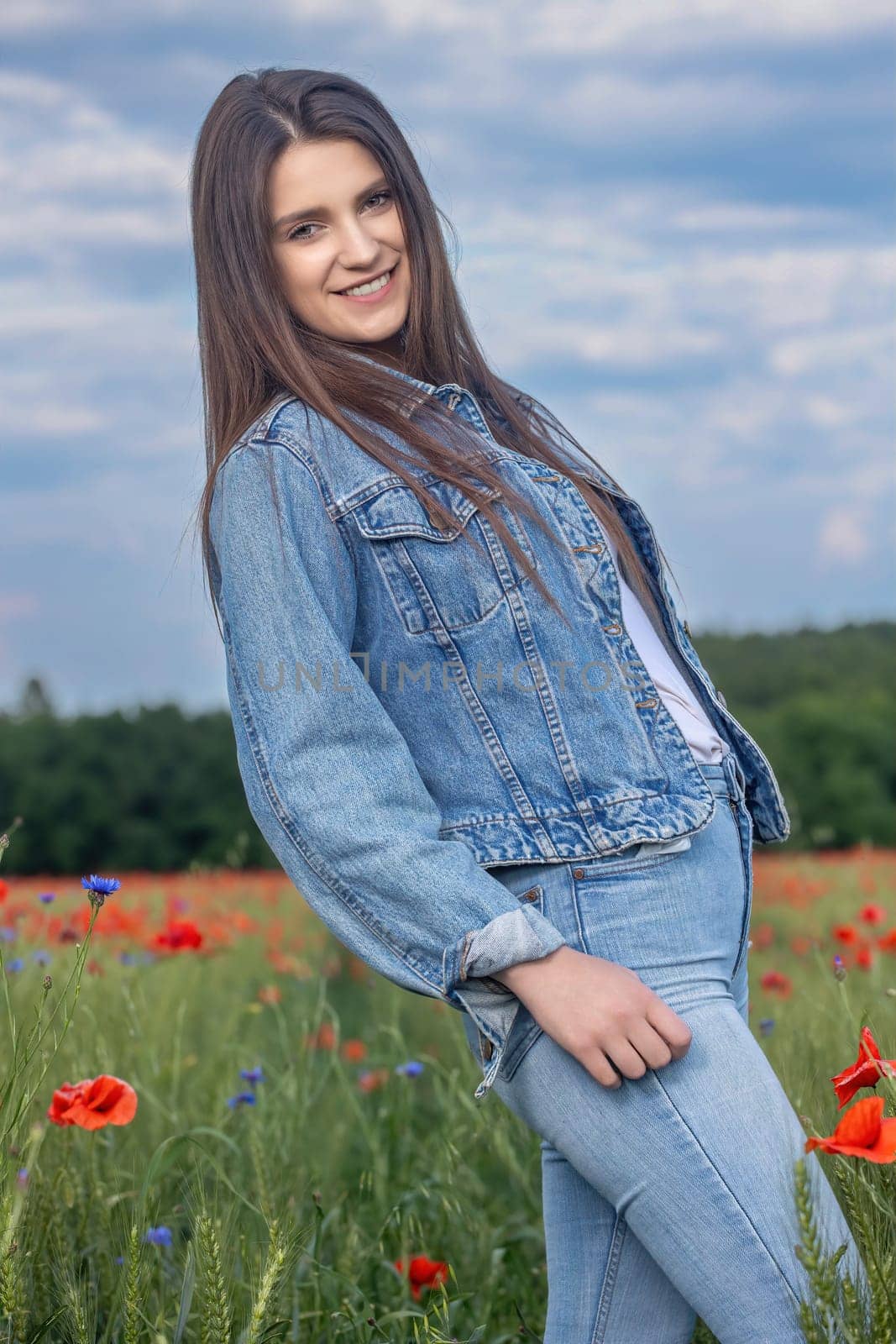 Smiling long haired  brunette girl dressed in blue jeans is posing in red poppies meadow. Vertically.