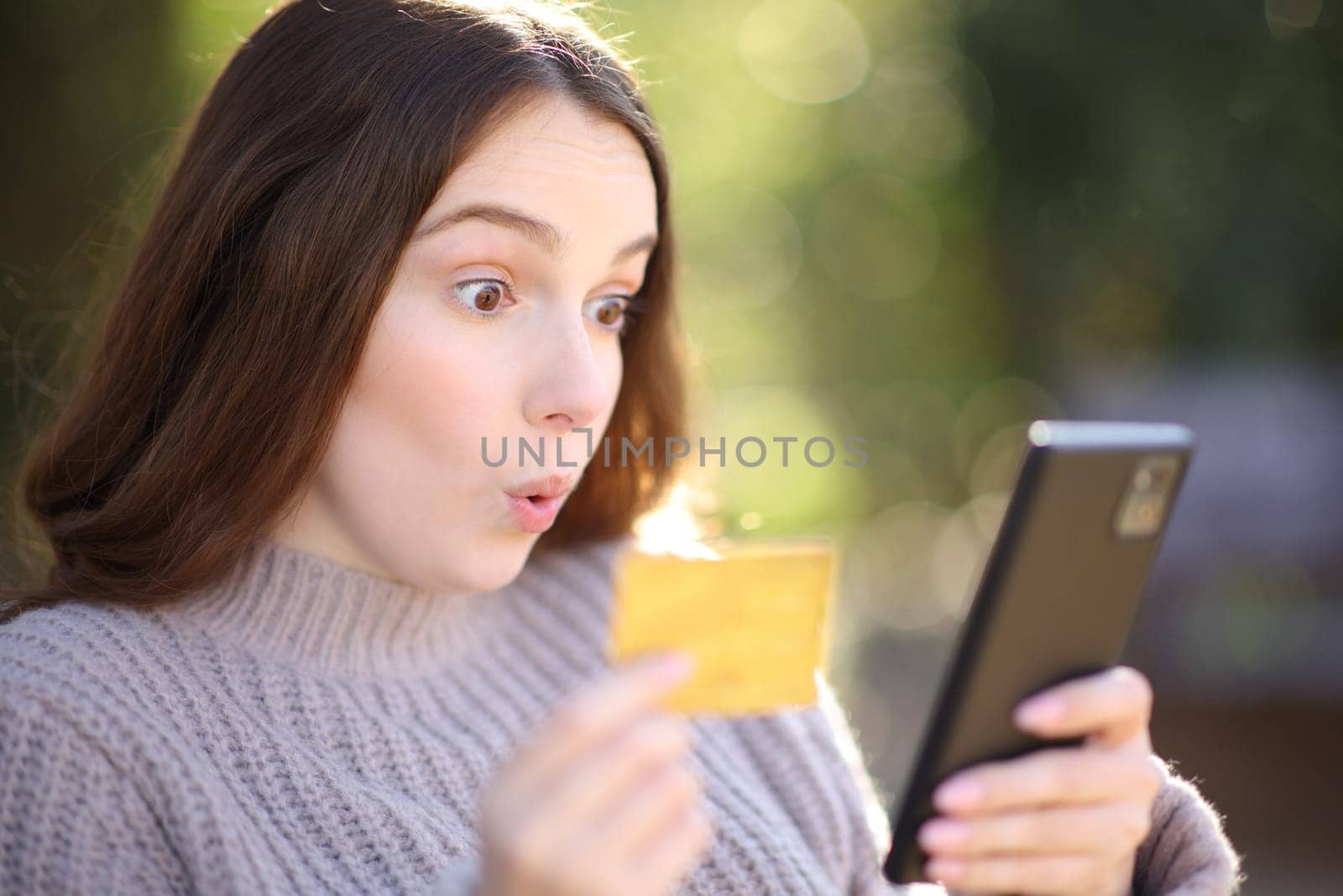 Surprised woman buying online with phone and credit card in a park