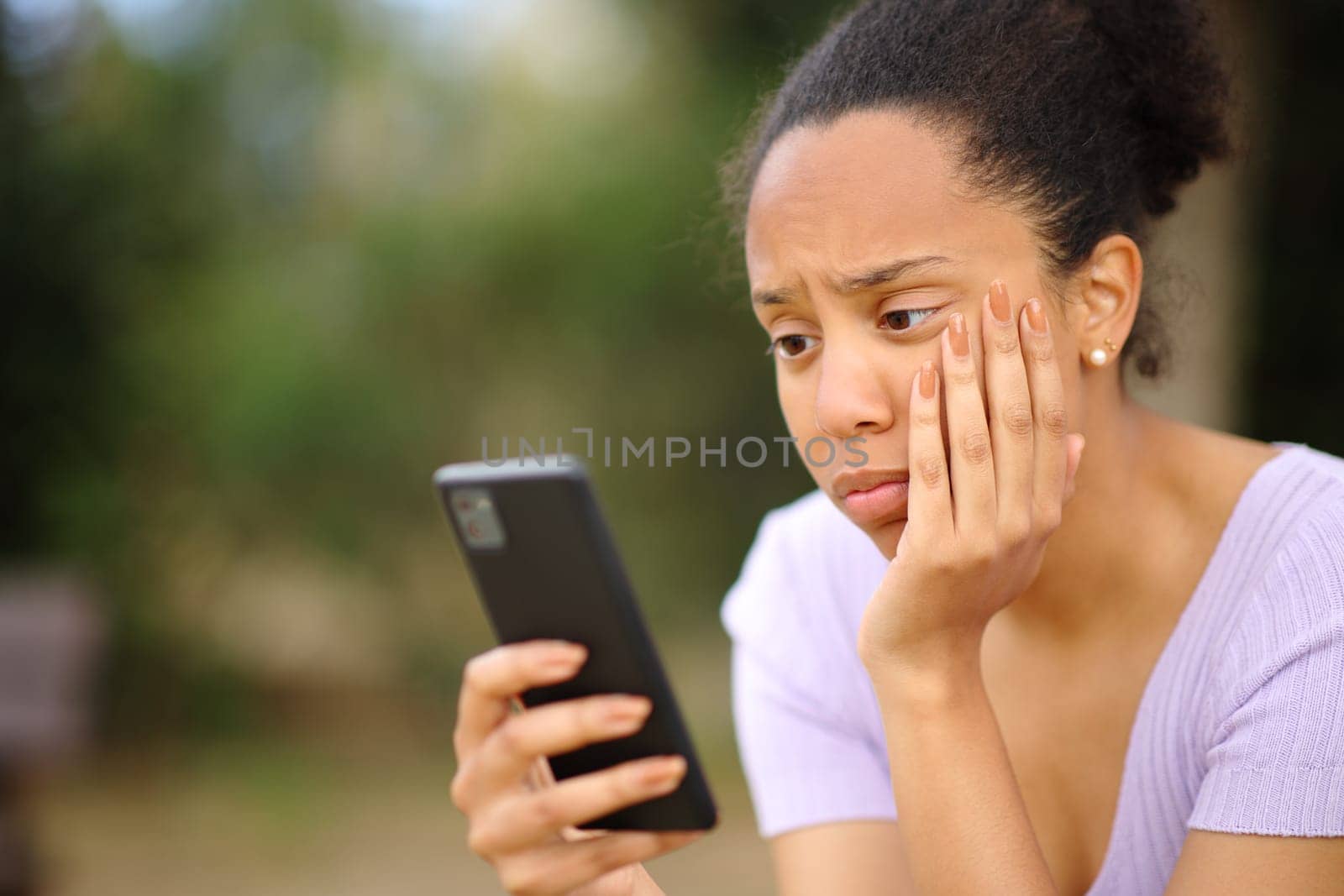 Worried black female checking cell phone sitting in a park