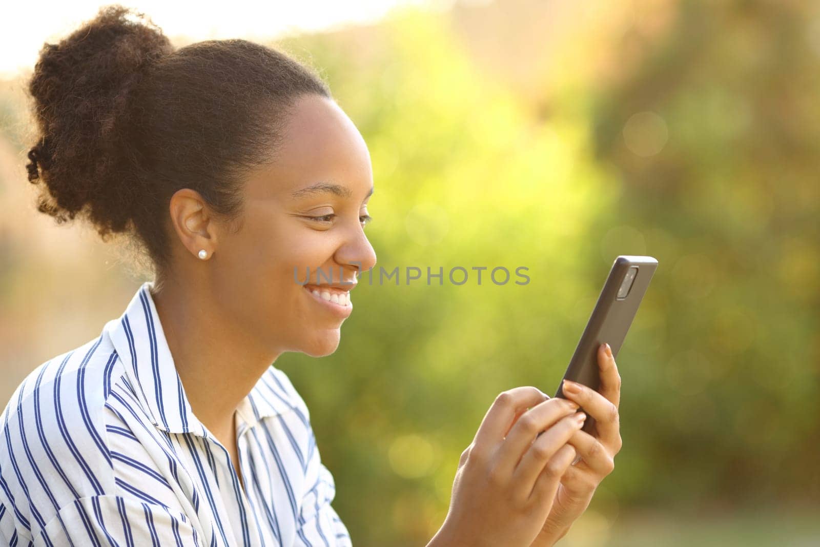 Happy black woman using cell phone sitting in a green park
