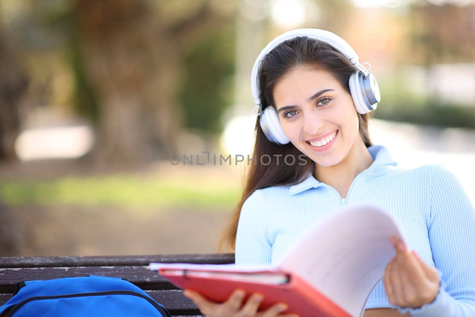 Happy student posing looking at you holding notebook and wearing headphone in a park or campus