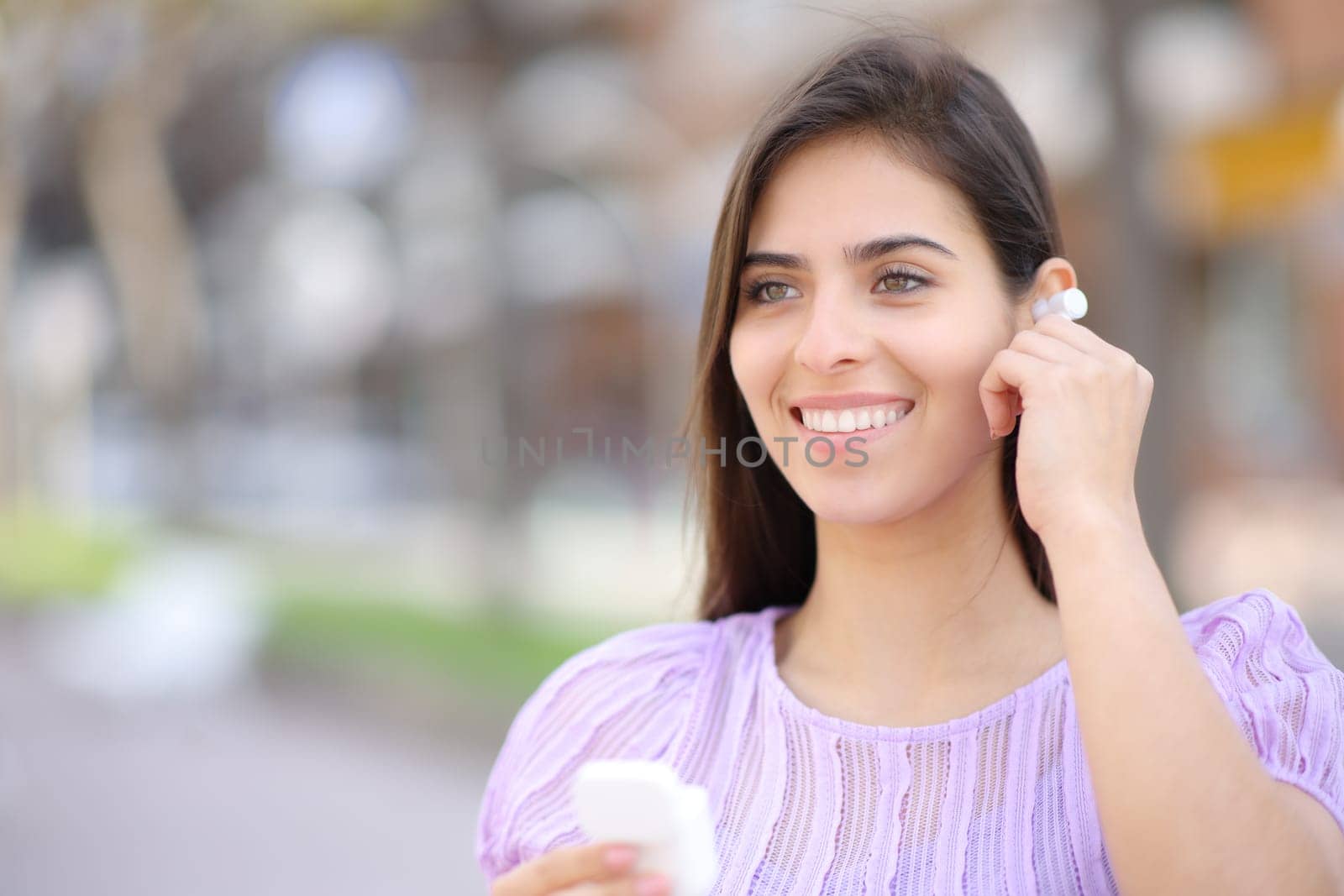 Happy woman putting earbud standing in the street looking away