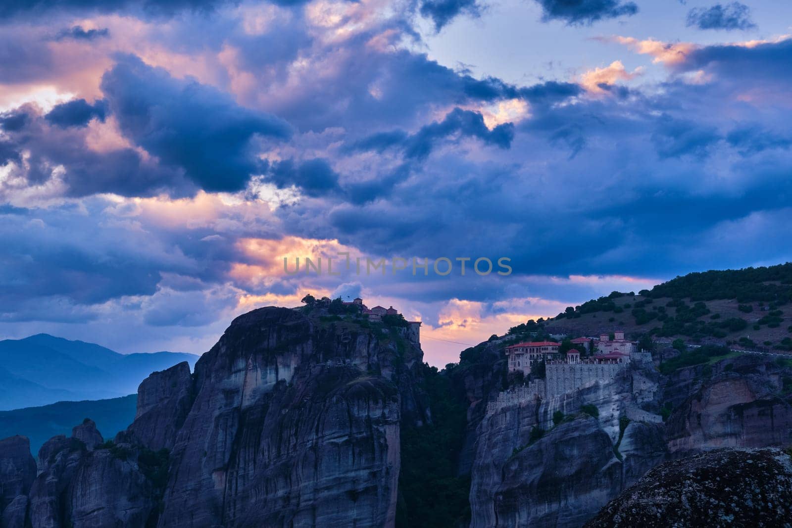 Twilight over clifftop Varlaam and Great Meteoron monasteries in Meteora, Greece. Underexposed, impressive sunset sky with clouds and sun rays. UNESCO World Heritage.
