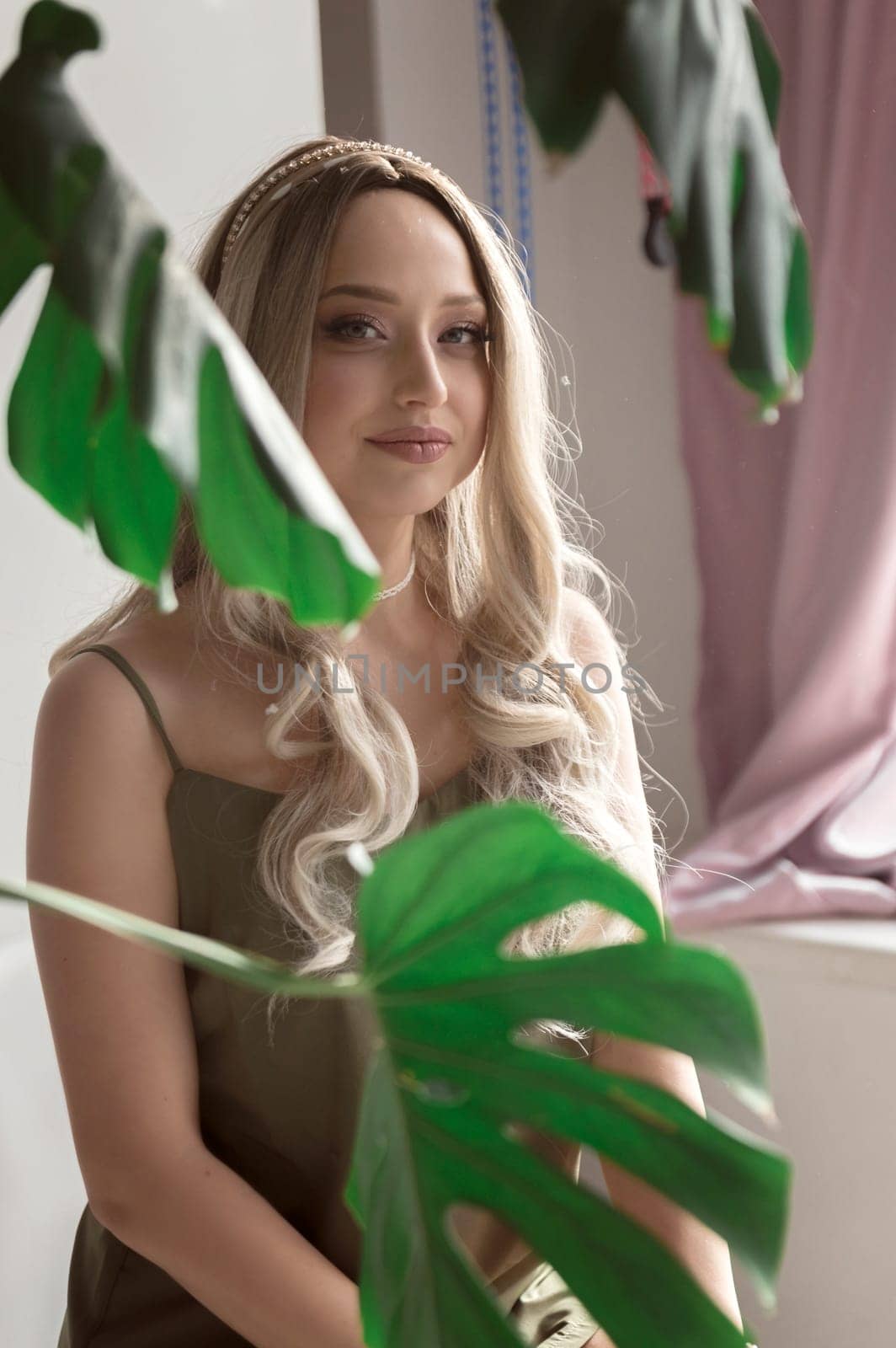 close-up of graceful blonde girl in thin dress sits on chair in minimalistic interior. girl has long light hair. she looks at camera through green monstera leaves. light wall behind her. green dress