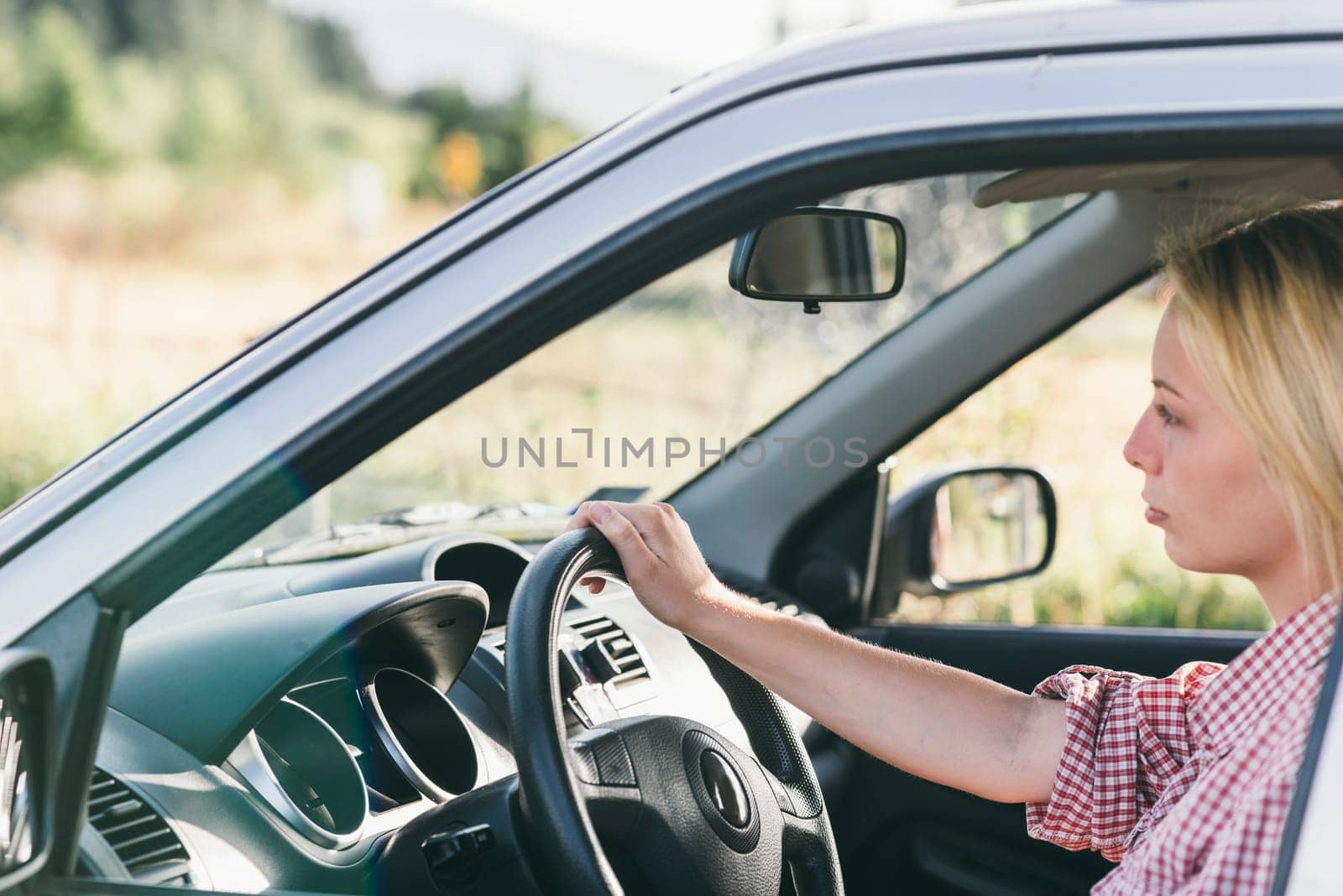 Young caucasian blond woman traveling by car and having fun on her summer holiday journey. Happy girl driving car on countryside road. Roadtrip vacation.