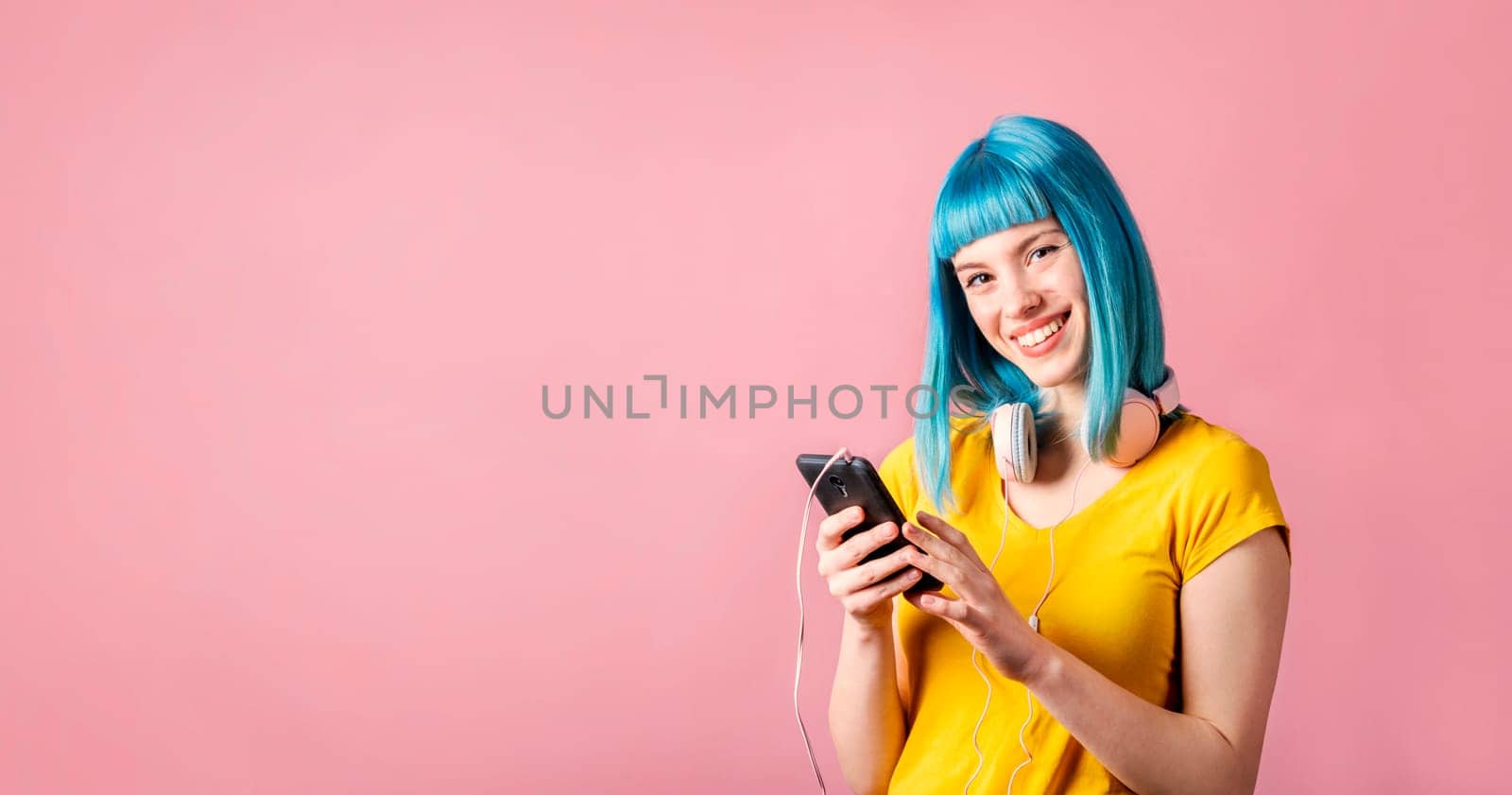 Trendy young woman with blue dyed hair holding smartphone and listen to music. Hipster girl with headphones over neon pastel background with copyspace.