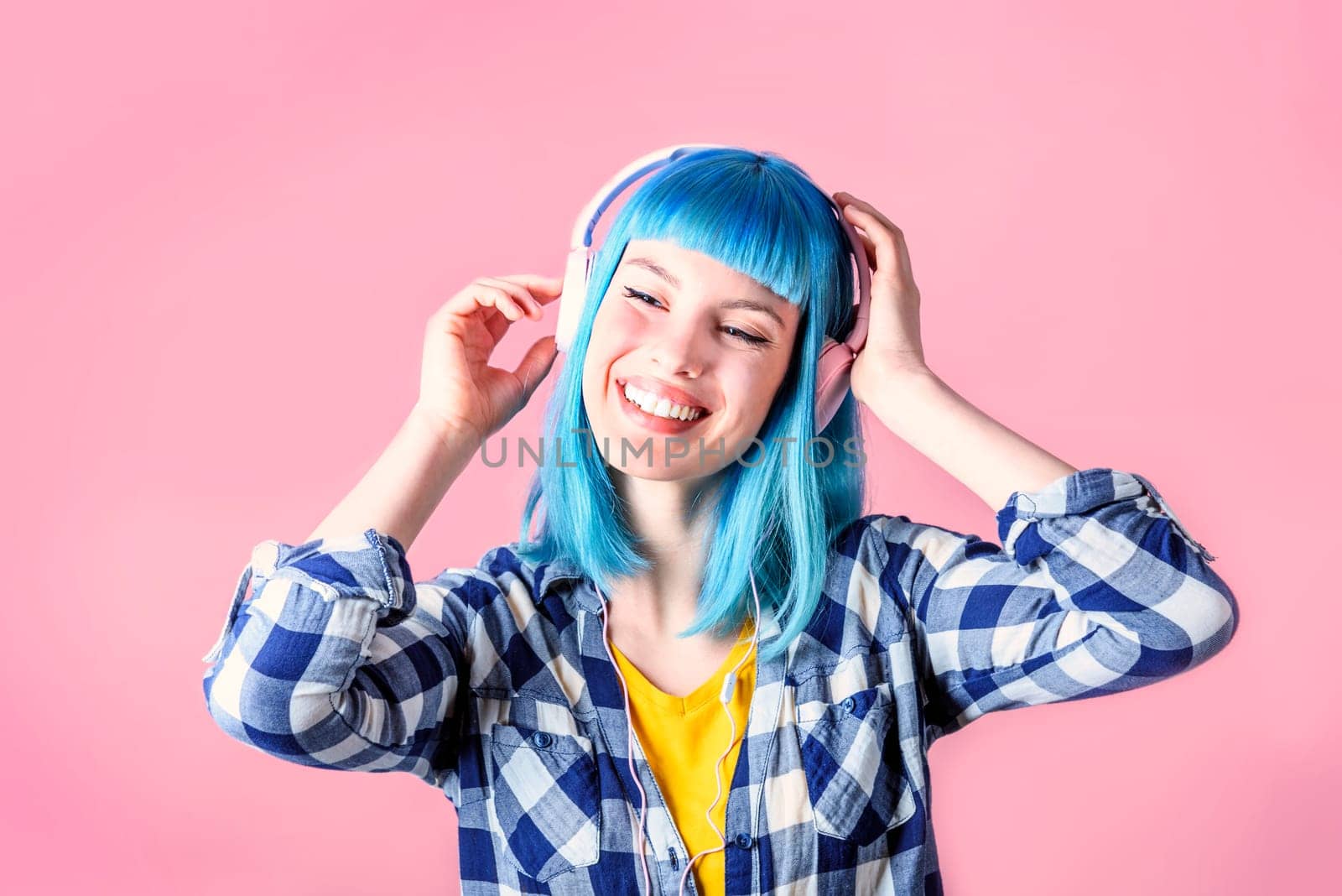 Trendy young woman with blue dyed listen to music and dancing. Hipster girl with headphones over neon pastel background.