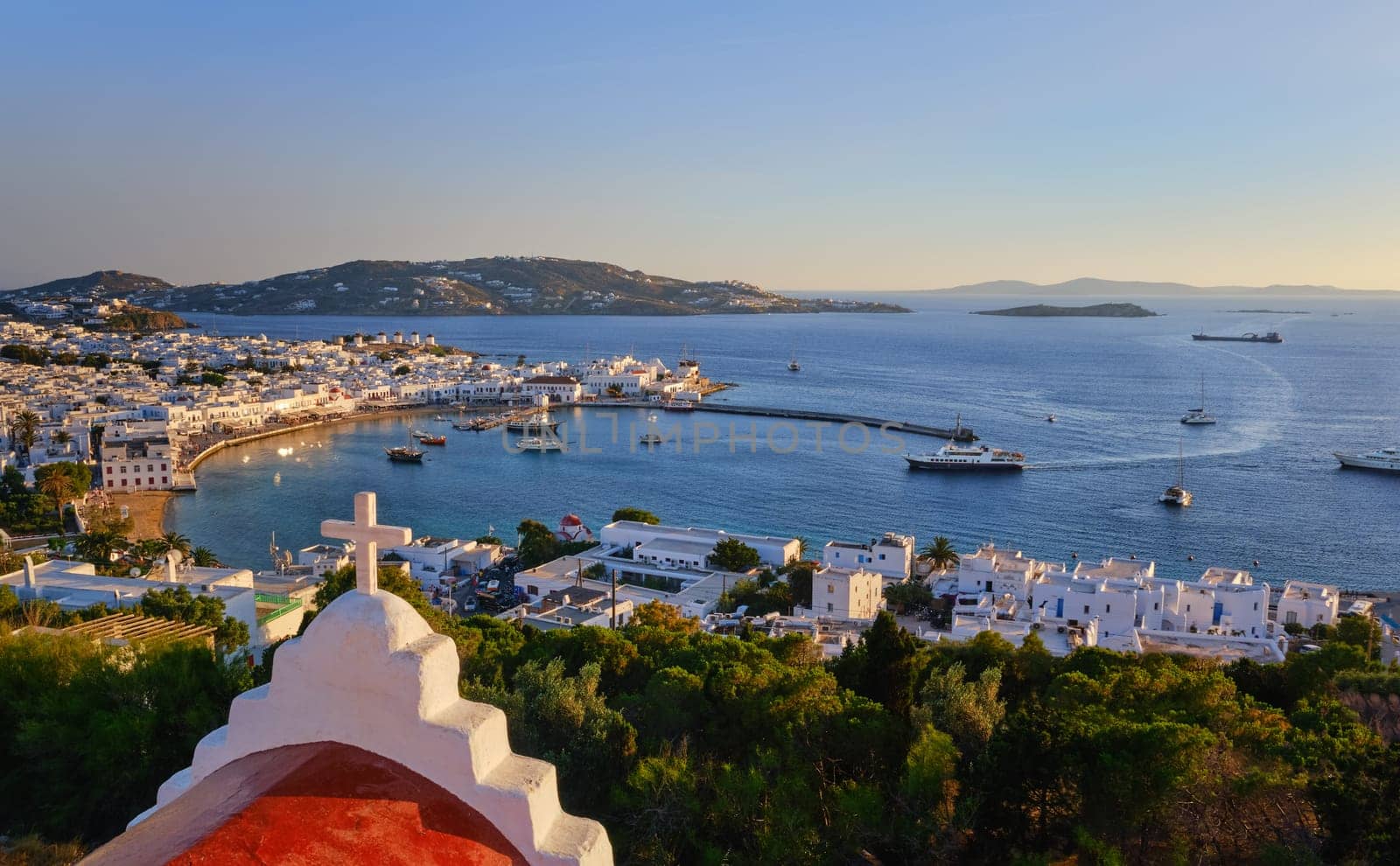 Beautiful sunset view over Chora, main town of Mykonos, Cyclades, Greece from red roof top of a Greek Orthodox church. Golden hour, harbor, ships, cruises arrive at port, whitewashed houses, distant islands in haze. Vacation, Mediterranean lifestyle