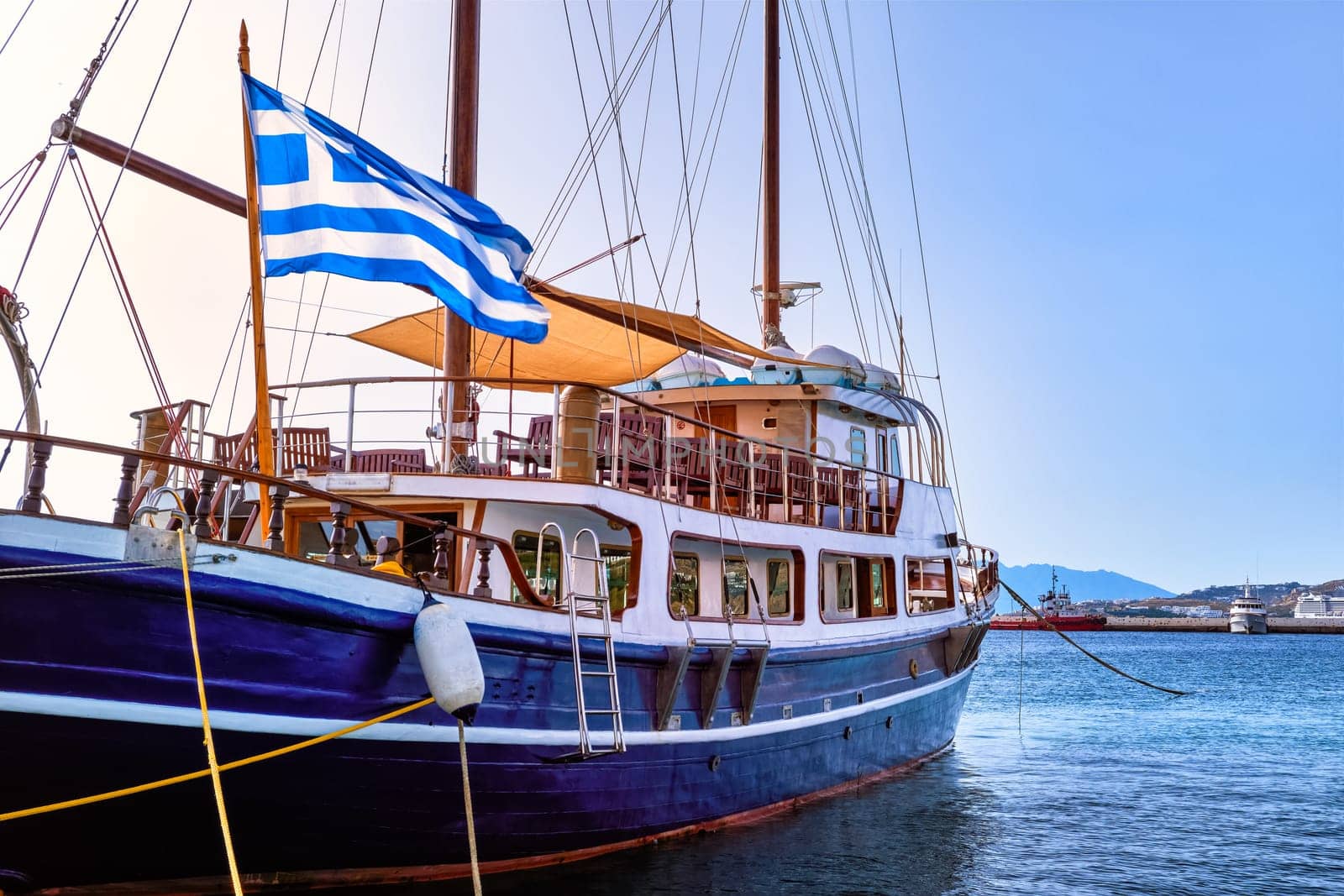 Schooner or sailboat under Greek flag moored at jetty of typical Greek island. Beautiful whitewashed houses on hills and by waterfront of harbor. Summer day sunshine, vacations and adventures. Mykonos, Greece.