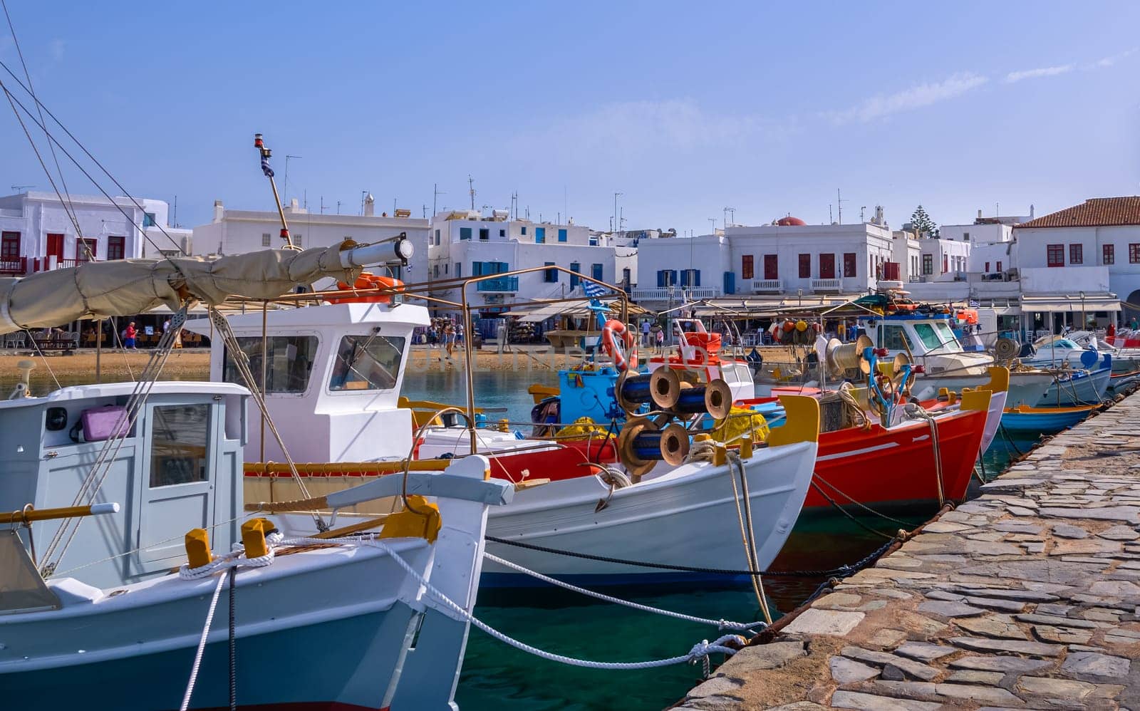 Beautiful summer day and sunshine in typical marina of Greek island. Fishing boats moored at jetty. Whitewashed houses by waterfront. Restaurants and shops. Mediterranean lifestyle, vacations. Fishing boats in foreground. Mykonos, Cyclades, Greece.