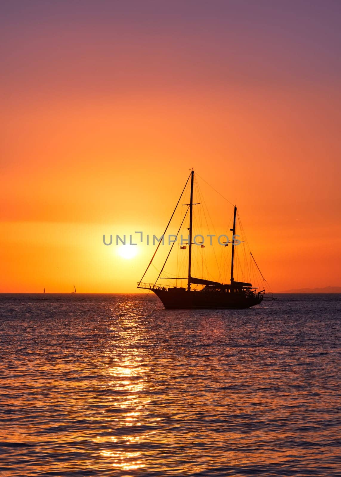 Silhouette of sailing boat with sails down against sun at sunset, sun glare on sea waters. Romantic seascape. Vertical shot. Holiday lifestyle landscape with skyline sailboat. Yachting tourism. Romantic trip on yacht during sea sunset
