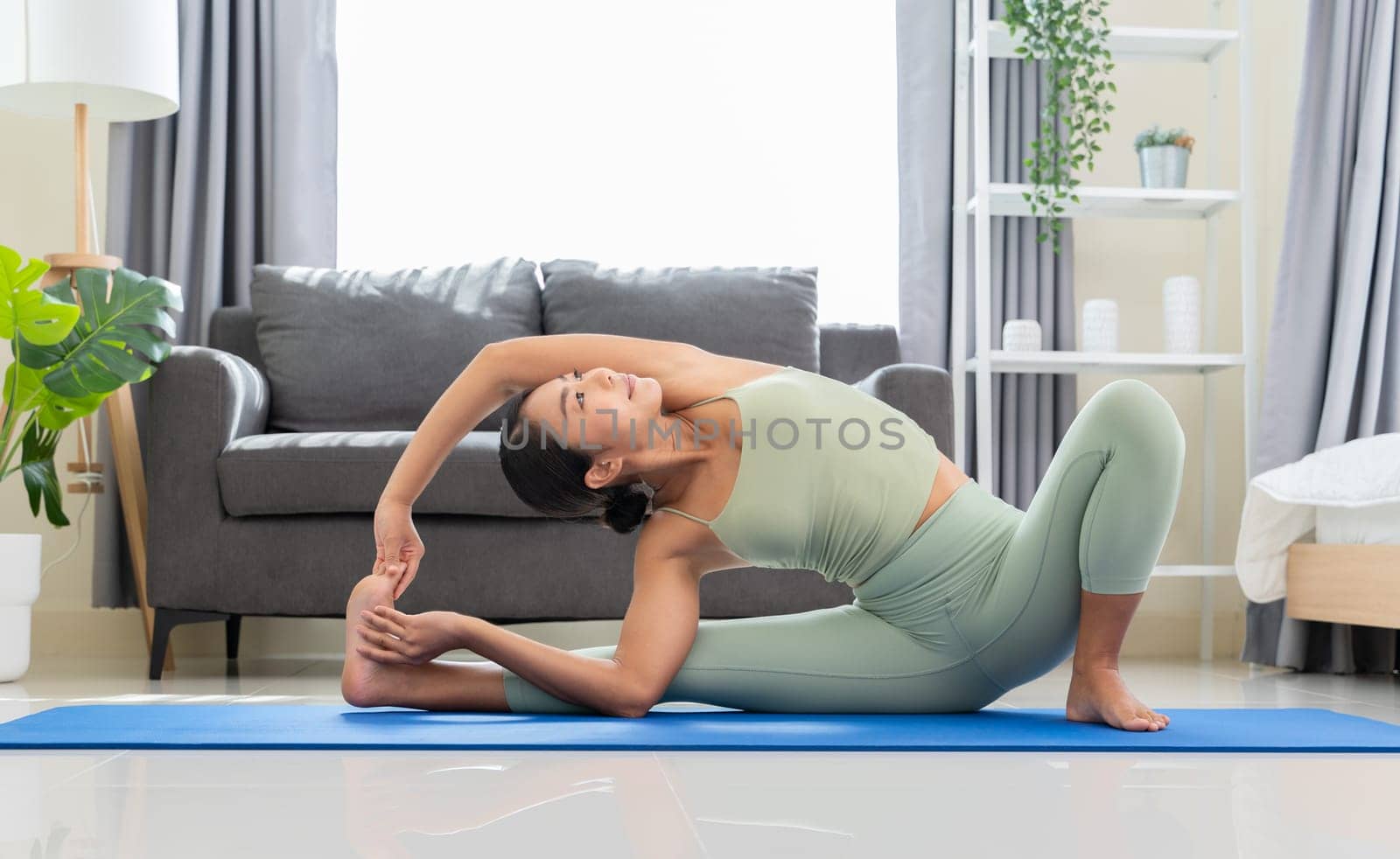 Young woman practicing yoga, sitting in Parivrtta Janusirsasana exercise or Revolved Head to Knee Pose on blue mat in bedroom. Healthy life and fitness training workout at home concept