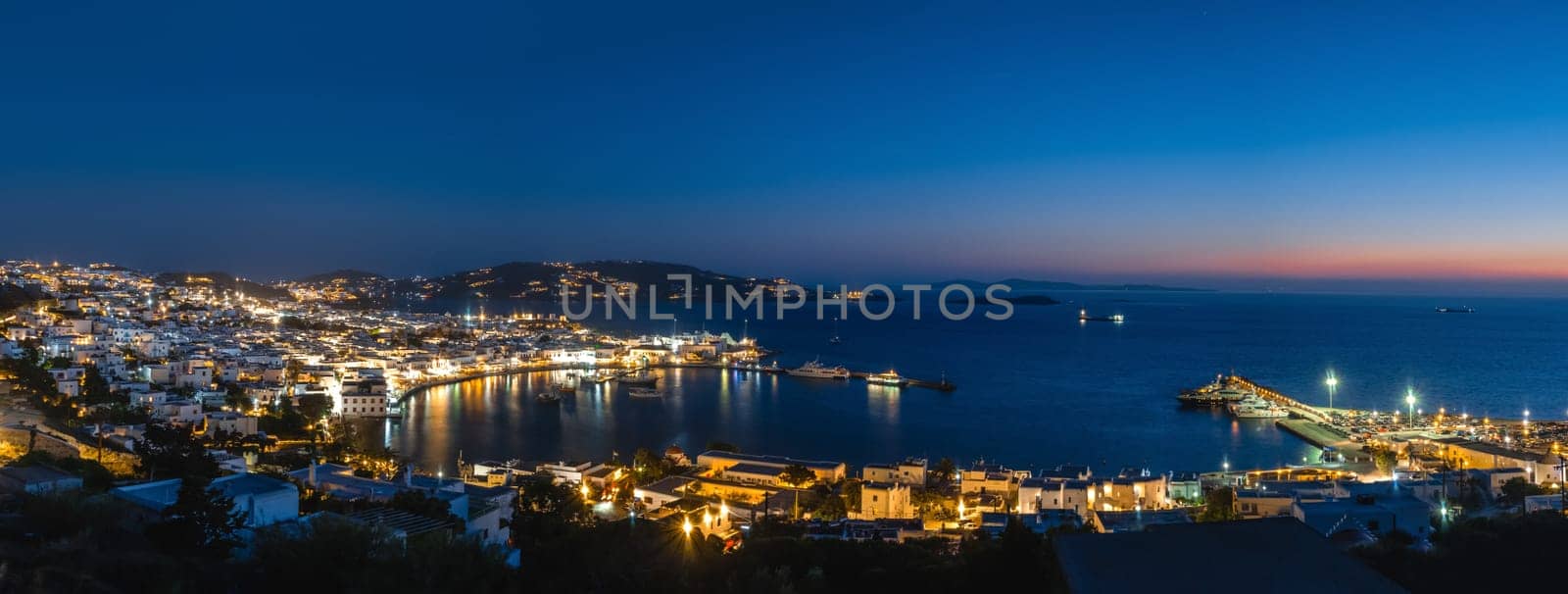 Iconic night panorama over Chora, main town of Mykonos, Cyclades, Greece. Harbor, ships, cruises, whitewashed houses and distant islands in hazy background. Town lights up. Vacations, leisure, nightlife, Mediterranean lifestyle