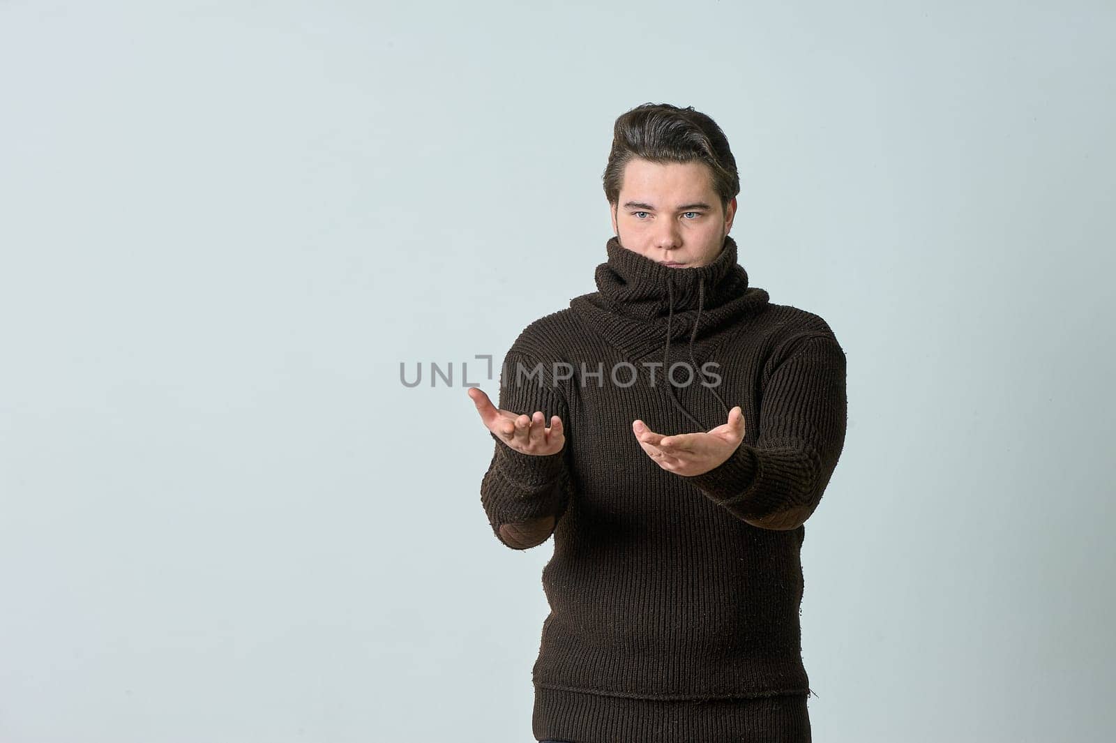 A man on a gray background in a brown sweater showing emotions, shows his hands. High quality photo