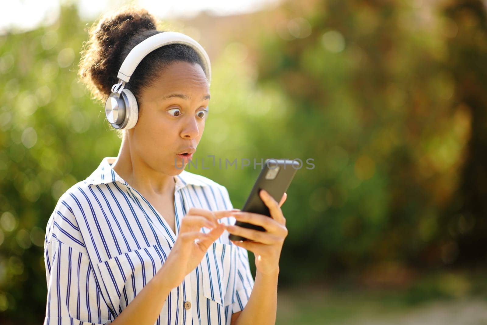 Surprised black woman wearing headphone listening to music on phone in a park