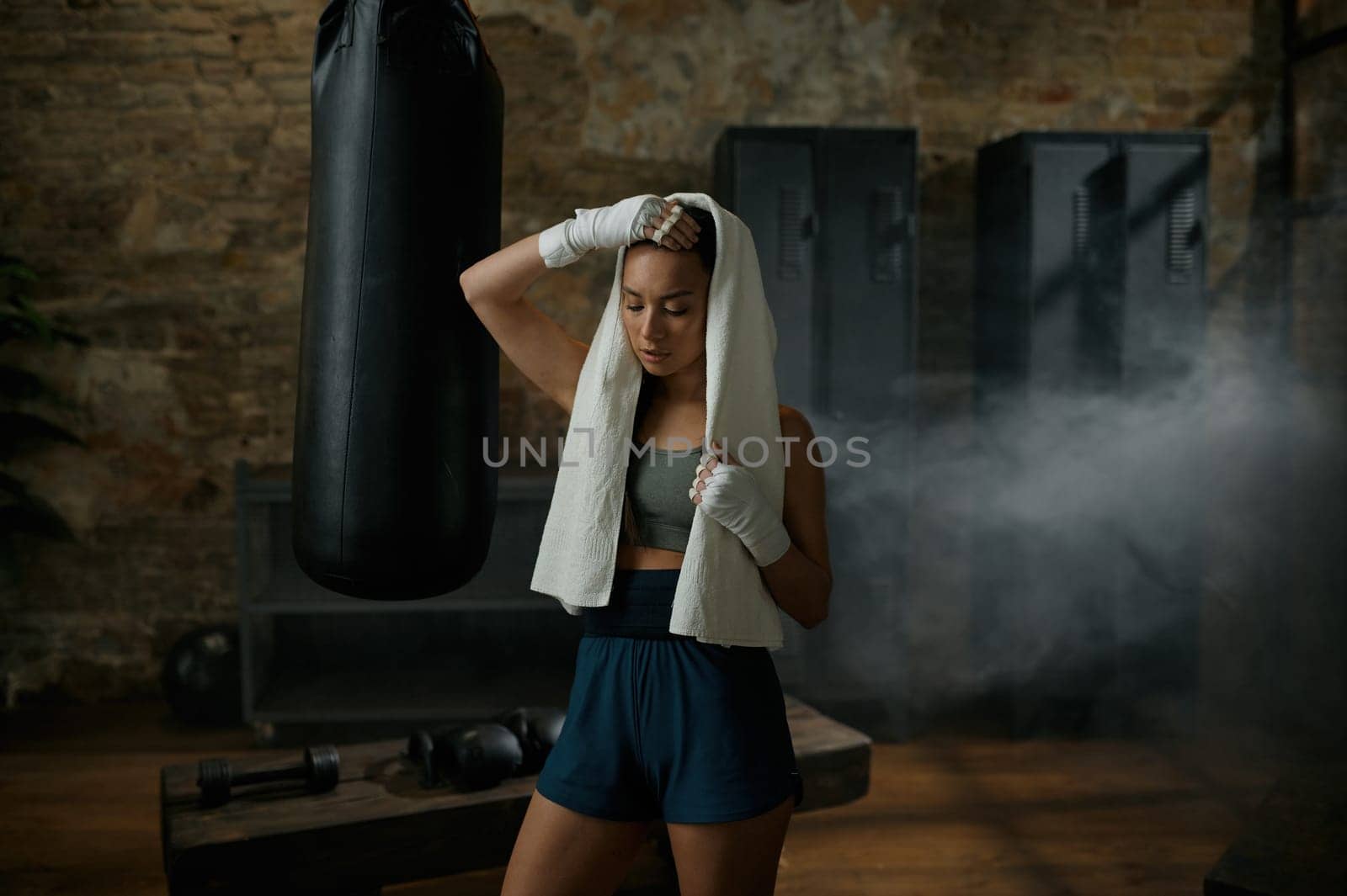 Tired sporty attractive woman boxer wearing towel on head and kickboxing bandages standing at gym take break after hard workout