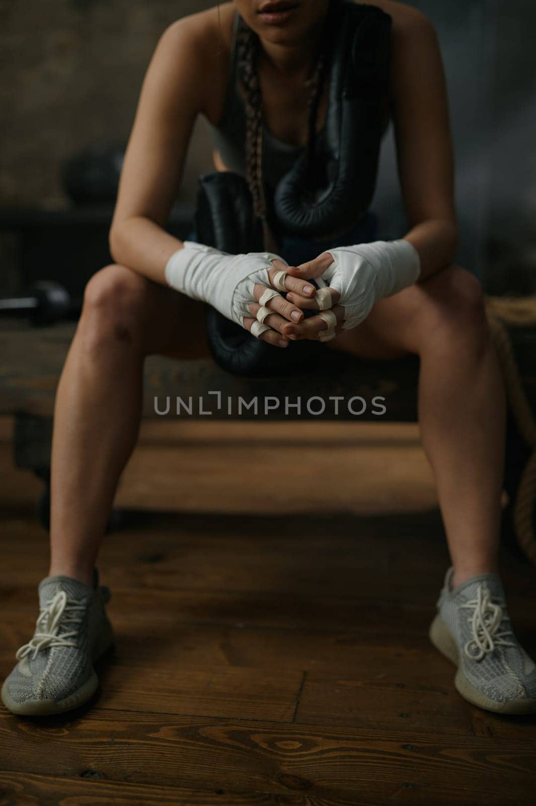 Crop shot of young sportive female boxer in boxing bandage sitting on bench over loft gym interior. Kickboxing training club for woman