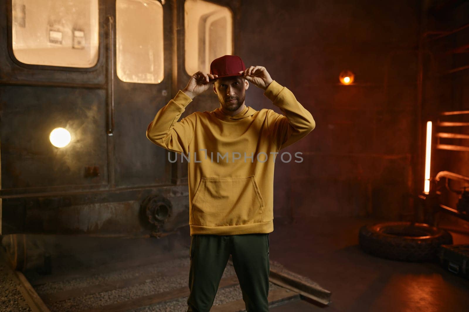 Portrait of man brake dancer touching his cap. B-boy posing for camera at studio with loft interior design and train on background